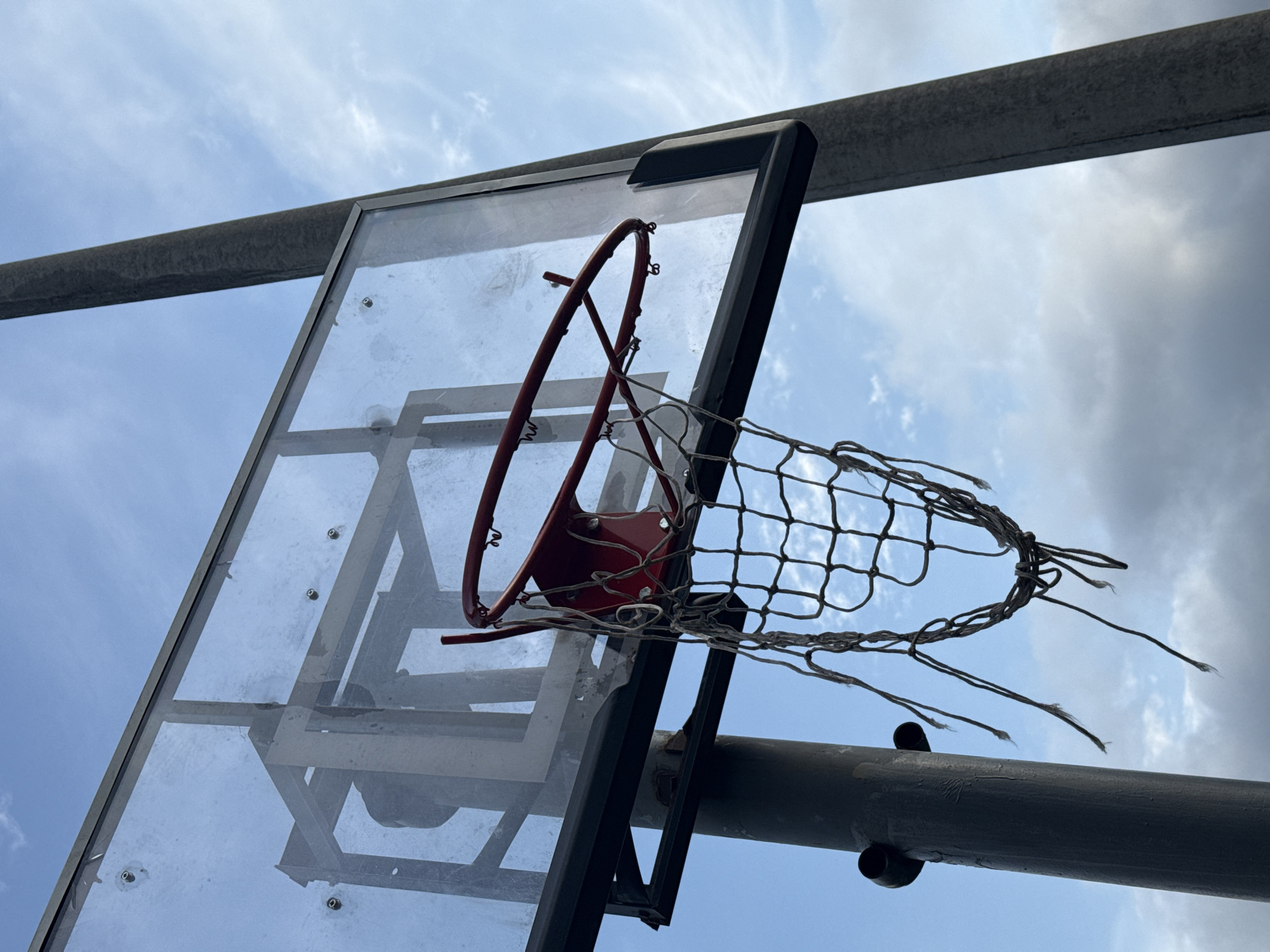 An upward-angle shot captures a basketball hoop with a transparent backboard against a cloudy sky. The basketball hoop, a red metal ring, is worn and detached in places, with a net that is tattered and frayed, with several strings hanging loosely. The transparent backboard has scuffs and smudges, and a metal frame supports it. A thick, gray pipe runs horizontally across the top of the frame. The lighting suggests it is daytime, with diffused sunlight breaking through the clouds, casting a soft glow on the scene. The overall impression is one of a well-used outdoor basketball court, likely in a park or recreational area, subject to the elements. There is no visible text in the image.