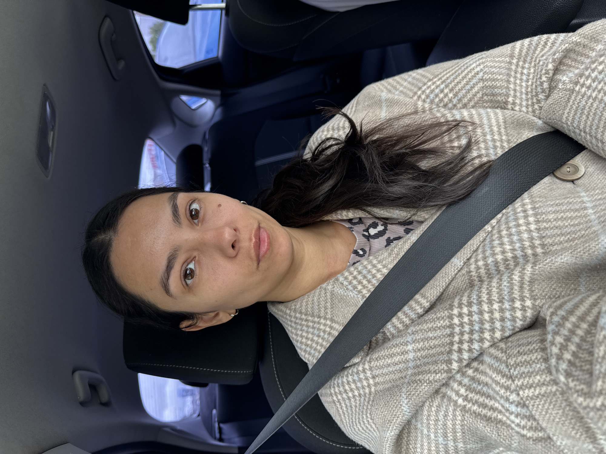 A woman is in the driver's seat of a car. She is looking directly at the camera with a neutral expression. She has long, dark hair, brown eyes, and light skin. She is wearing a beige and white plaid jacket over a grey leopard-print top. A black seatbelt is visible across her chest. The interior of the car is dark, with black seats and dark blue upholstery. The background shows the car's interior, including the headrests and windows. The image appears to be a selfie taken inside the car.