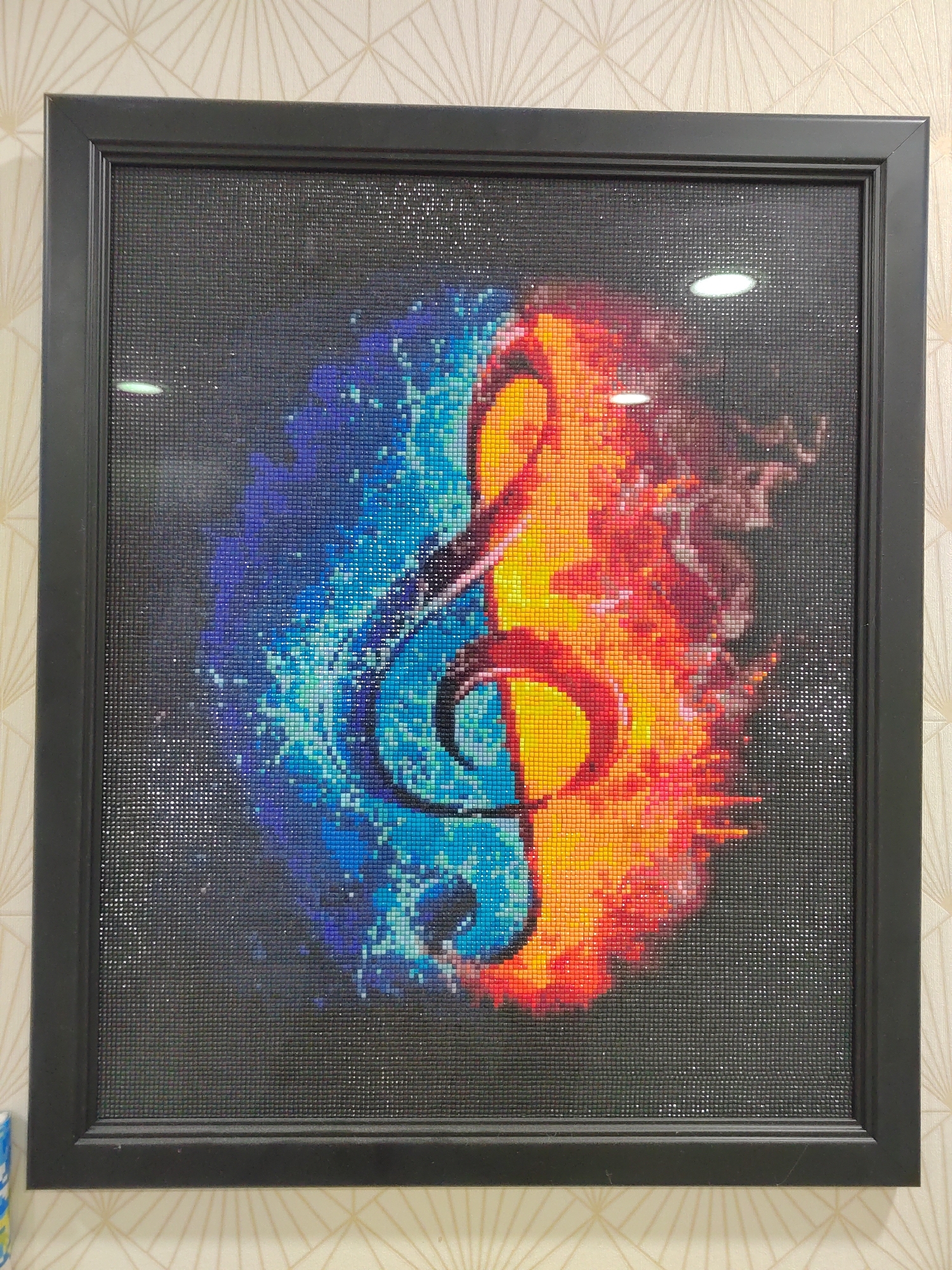 A framed picture hangs on a wall with patterned wallpaper. The picture, with a black frame, depicts a treble clef. The left side of the image surrounding the clef is colored in shades of blue, while the right side is colored in shades of red and orange, possibly representing water and fire. The background is dark and covered with tiny sparkling squares. The location is Podgorica, Montenegro.