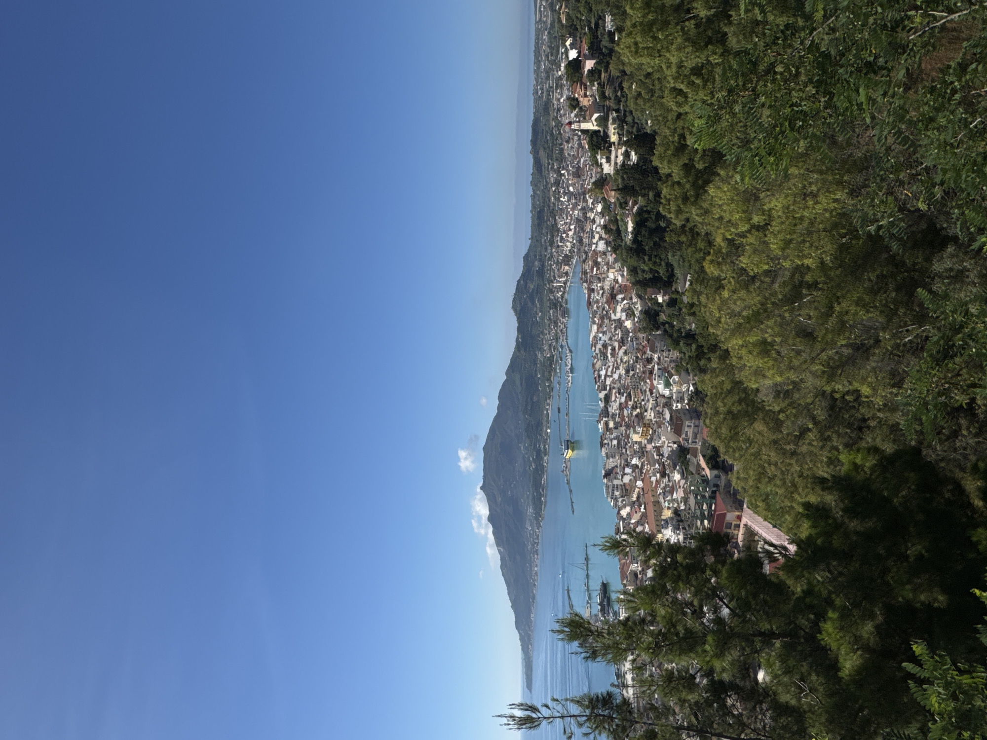 This is a high-angle, wide shot of the city of Zakynthos on the island of Zakynthos, Greece, on a clear, sunny day. The foreground is dominated by lush green trees and pine trees, with a pink pathway visible winding through them. In the midground, the city of Zakynthos unfolds with its dense network of buildings featuring red-tiled roofs. A prominent church with a red dome and a white bell tower stands out amongst the buildings. The city borders a tranquil, turquoise bay filled with boats of various sizes, including sailboats and a large yellow ferry. Across the bay, a mountainous landscape stretches into the distance, with smaller settlements dotting its slopes. The sky is a vibrant, cloudless blue.