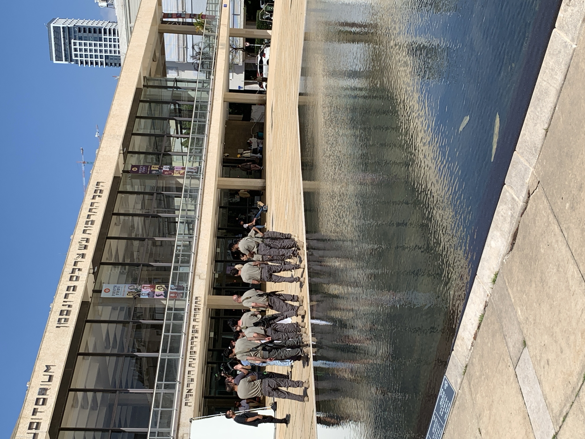 The image depicts a scene in Tel Aviv, Israel, near the Tel Aviv Performing Arts Center, also known as the Israeli Opera House. Several individuals dressed in khaki shirts and dark tactical pants with equipment strapped to their chests are standing along the edge of a shallow, rectangular body of water, possibly a reflecting pool or fountain. The water reflects the building's facade. A man is standing to the left, dressed in black. The building itself is a modern architectural structure with large glass windows and a beige exterior, and the words "המשכן לאמנויות הבמה" are inscribed vertically on the building's left side, meaning "The Performing Arts Center" in Hebrew. A tall, white high-rise building is visible in the background under a clear blue sky.