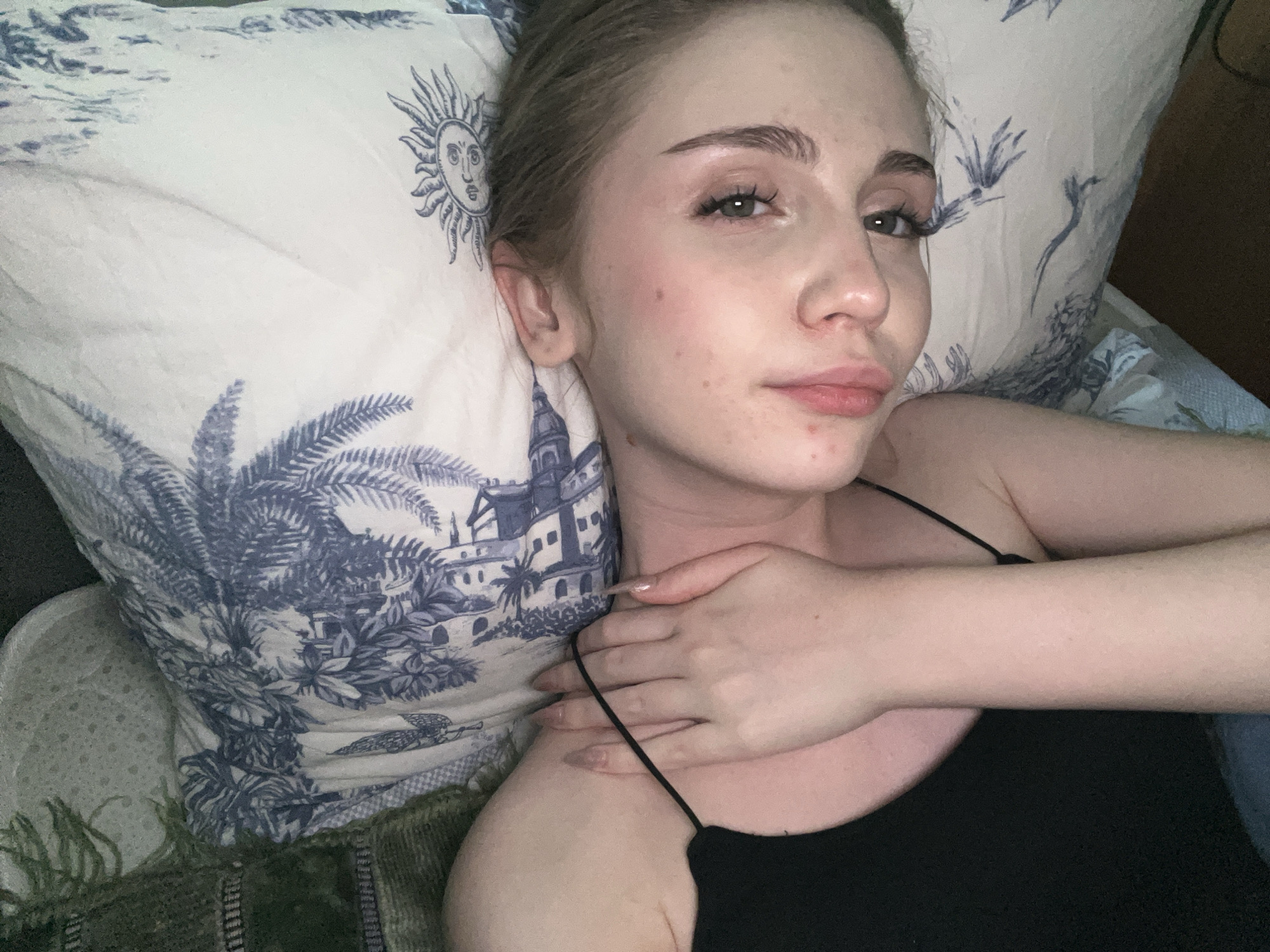 A young woman is lying on a bed with a pillow that has a blue and white pattern of palm trees and a sun. She is wearing a black tank top. Her right hand is resting on her neck. The room is dimly lit. No information is available regarding the city of Ashkadar, Russia.
