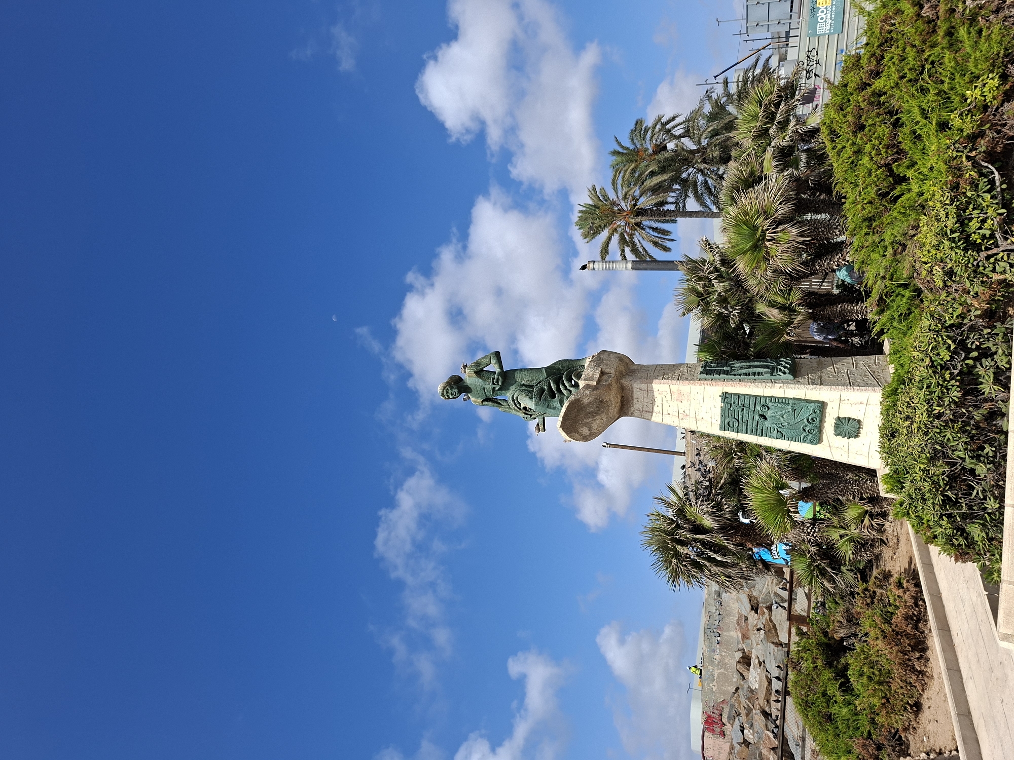 The image shows a large bronze statue of a figure standing on a monumental base. The statue is located in a public area with palm trees and lush green vegetation. The sky is blue with scattered white clouds, and there is a crescent moon visible in the upper left quadrant of the sky. In the background, there are some industrial structures and a construction site with scaffolding.
The statue depicts a standing male figure with a determined expression, wearing a tunic. He is holding an object, possibly a spear or staff. The base of the monument is made of stone and features some decorative reliefs.
The location appears to be a waterfront or coastal area, as suggested by the palm trees and the open sky. The presence of industrial elements in the background might indicate a port or a working area near the coast. The overall impression is one of a monument situated in a public space, possibly a park or a promenade, offering views of the sea or the city.
The statue and its surroundings seem to be well-maintained, though the construction in the background suggests ongoing development or renovation in the area. There are no specific identifiable landmarks that pinpoint the exact location in Torrevieja, Spain, but the presence of palm trees and the clear blue sky are consistent with a Mediterranean climate.