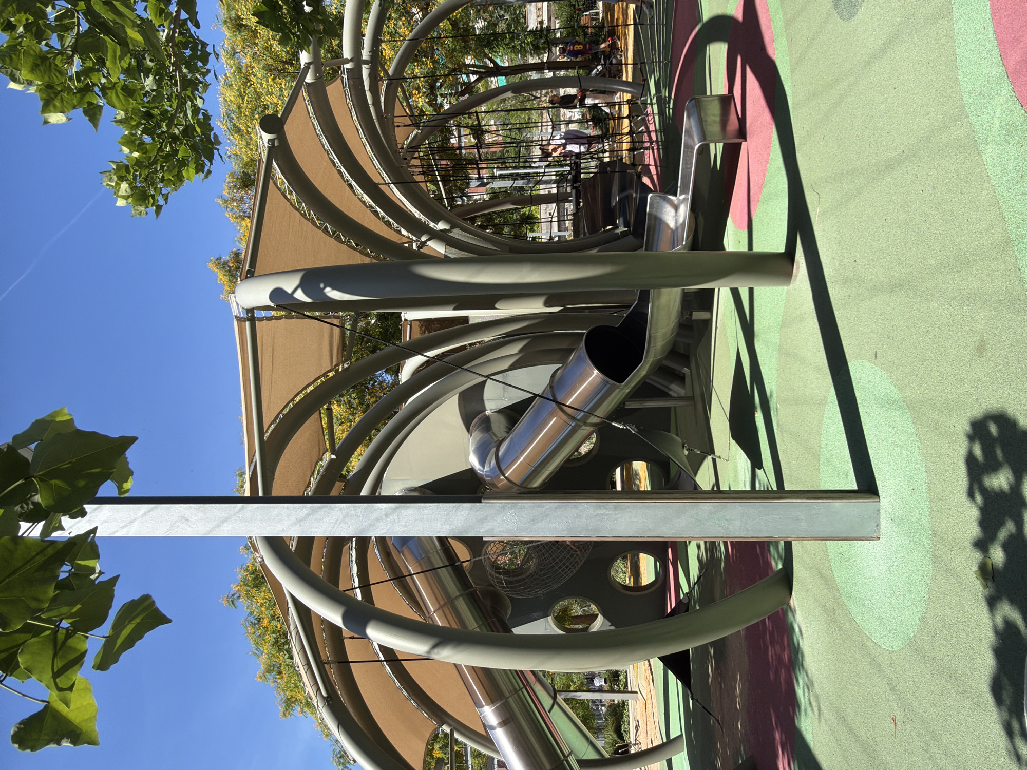 This is a sunlit, medium-angle exterior shot of a modern playground structure in Barcelona, Spain. The structure, made of gray metal supports and tan fabric canopies, features curved tubes, slides, and rope netting. Two silver slides are visible, one descending towards the foreground and another curving into the structure. The playground surface is a multi-colored rubberized material in shades of green and pink. In the background, through the gaps in the structure, several people are visible, including adults and children, with some walking and others near what appears to be a stroller. The environment is characterized by a clear blue sky and lush green trees, some with yellow blossoms, suggesting a warm day. The overall impression is of a vibrant and well-maintained urban recreational space.