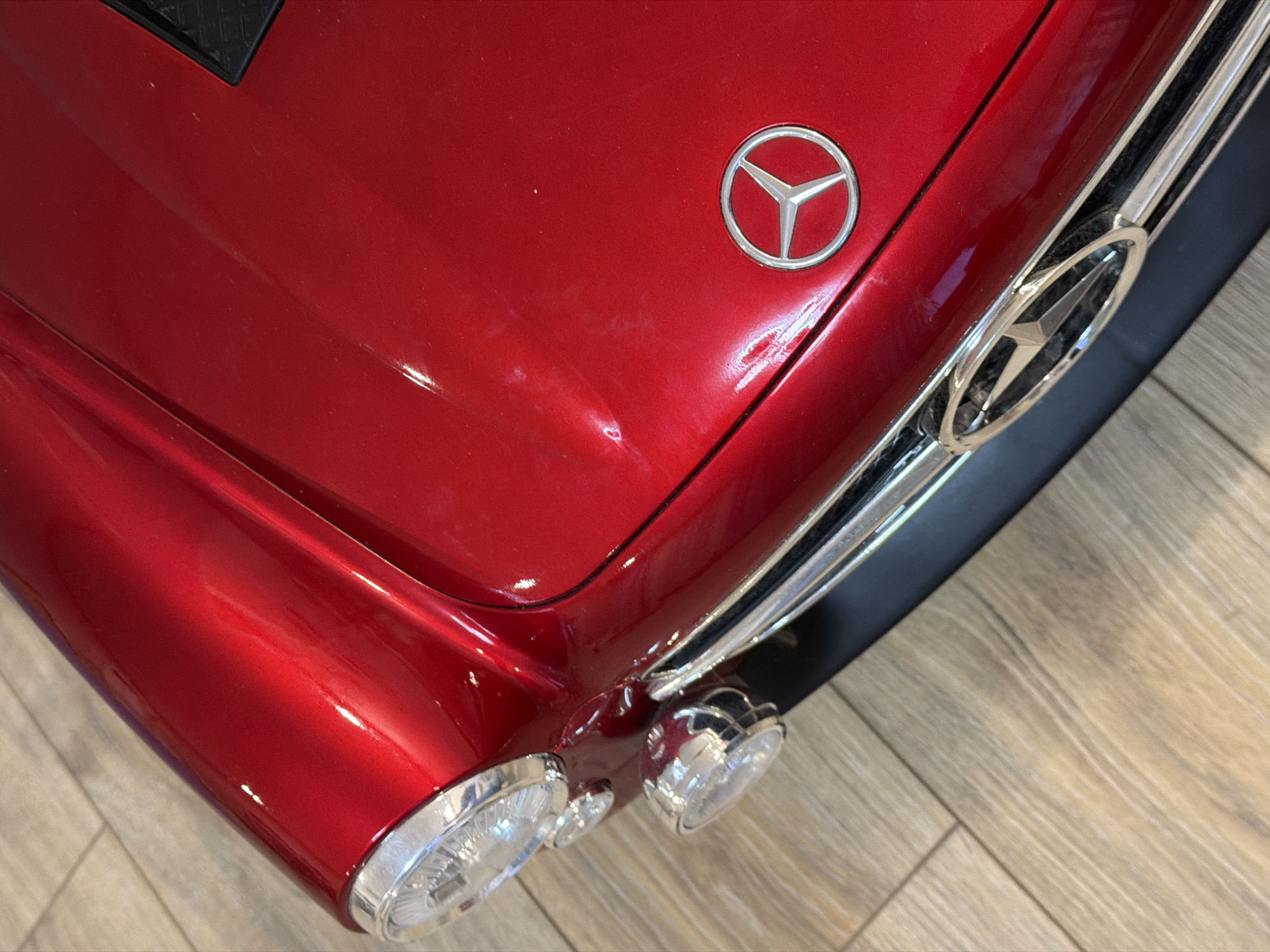 A high-angle, close-up shot shows the front of a red Mercedes-Benz car. The vehicle's hood displays the Mercedes-Benz logo. Chrome accents are visible on the grill, headlights, and around the larger logo. The car sits on a surface resembling light-colored wood flooring. The location is unspecified, and there is no indication that the photo was taken in Chaikovsky, Russia, nor is there any evidence of people or actions.