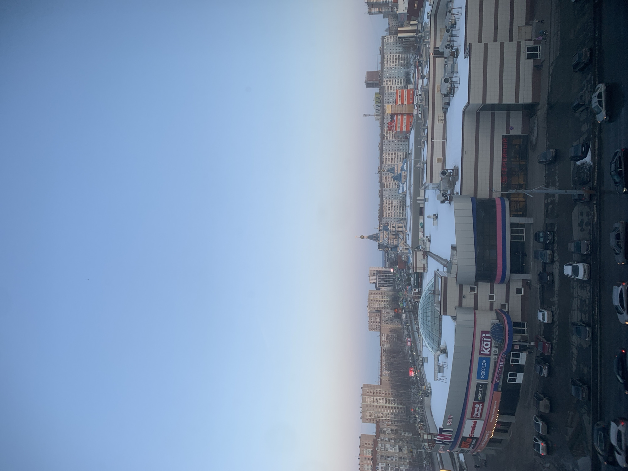 The image depicts an elevated view of a cityscape in Izhevsk, Russia, under winter conditions, likely during late afternoon or early evening. The prominent structure in the mid-ground is the "ТЦ ЦЕНТР" (TC Center) shopping mall, located at ulitsa Karla Marksa, 274.
The shopping mall is a multi-story building with a light-colored facade featuring horizontal beige, white, blue, and pink stripes. Its flat roof is covered with snow and includes a large central glass dome, along with several ventilation units and pipes. Numerous commercial signs are visible on its exterior, including "SOKOLOV", "kari", "Пятёрочка" (Pyaterochka), "М.ВИДЕО" (M.Video), "DNS", "Красное&Белое" (Krasnoe&Beloe), "СЕМЕЙНЫЙ" (Semeynyy), and "Эльдорадо" (Eldorado).
In front of the mall, a dark, wet asphalt parking lot contains approximately 20 to 30 parked passenger vehicles of various models and colors. A main road with visible vehicle traffic is situated to the left of the mall. A few indistinct human figures are present on sidewalks near the mall entrances.
In the background, beyond the shopping mall, numerous multi-story residential buildings of utilitarian design are visible. The distinctive St. Michael's Cathedral (Михайловский собор) is prominently featured in the central background, characterized by its blue onion domes adorned with gold stars, a golden main dome, and light-colored walls. Further in the distance, additional urban structures and a cellular tower are visible. Bare trees line some streets.
The sky is clear and cloudless, displaying a gradient from a medium blue overhead to a lighter, slightly pinkish hue near the horizon. Snow covers the rooftops of buildings, indicating cold temperatures. Roads appear wet and dark.