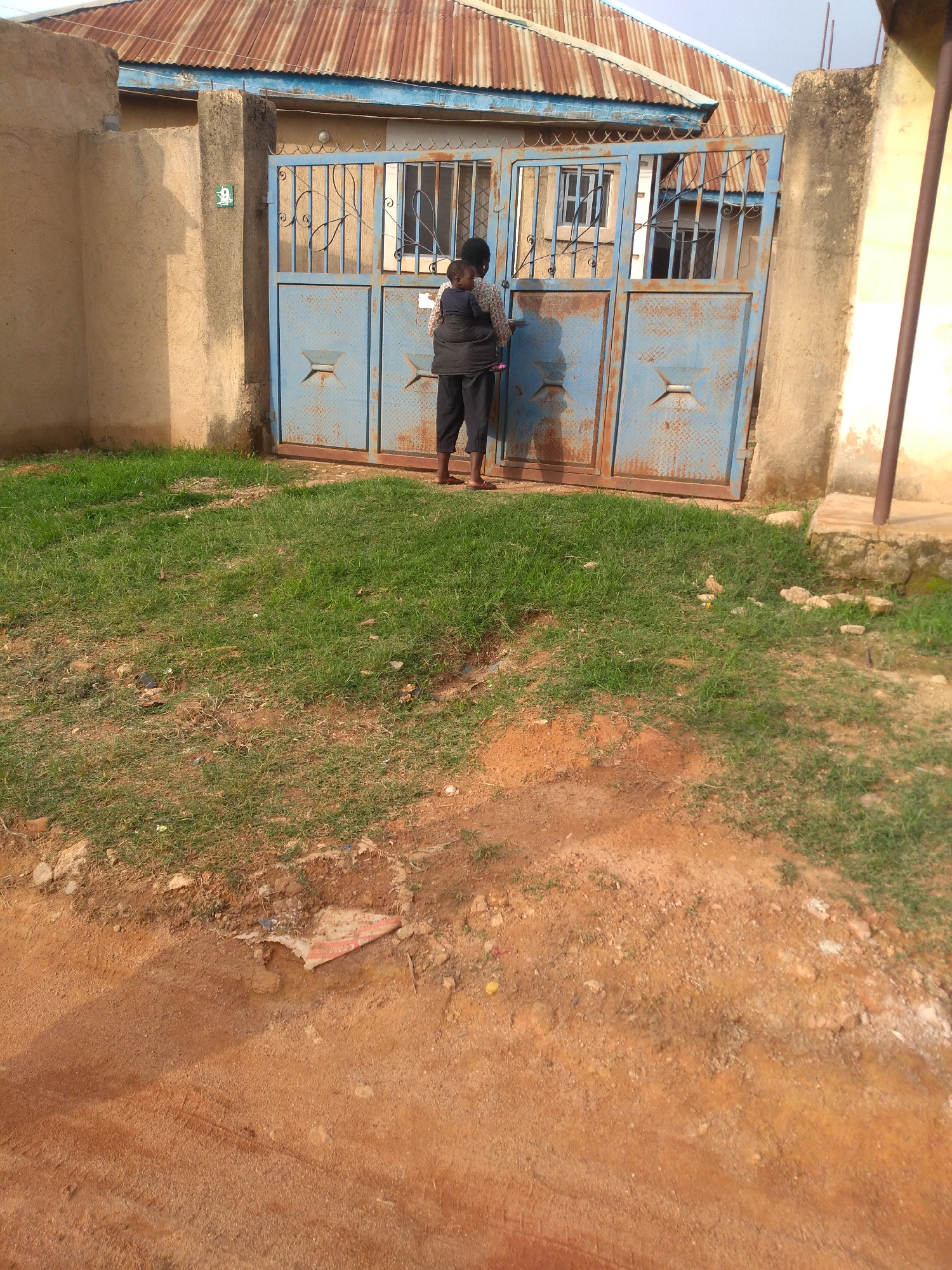In Jos, Nigeria, a person stands in front of a blue corrugated metal gate. The gate is weathered and shows signs of rust. Behind the gate, a building with a corrugated metal roof, painted in shades of orange and brown, is visible. The building has windows with metal bars. A grassy area is in front of the gate, with a dirt path leading away from it. The overall environment suggests a residential area.