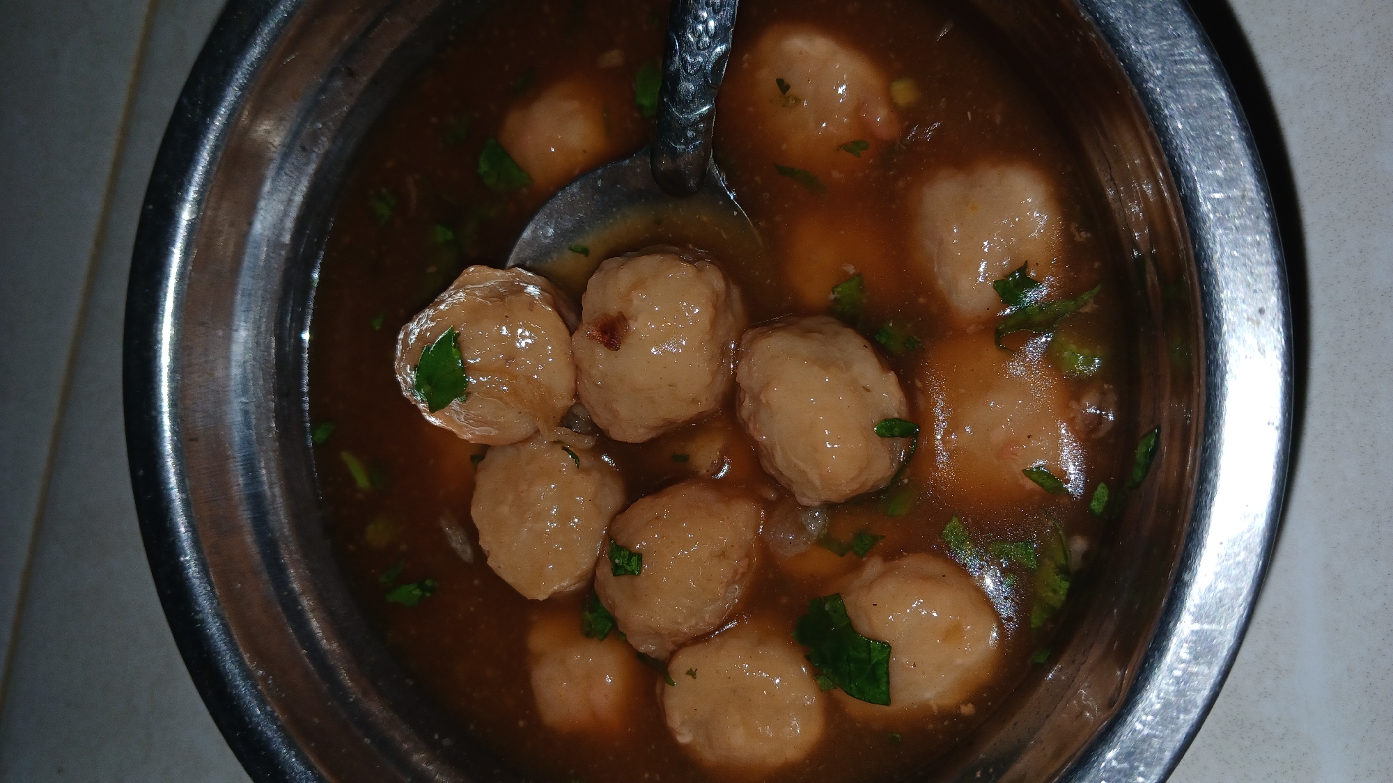 A metal bowl filled with meatballs in broth, garnished with green herbs, is partially lifted by a spoon. The setting appears to be indoors, likely a home in Lhokseumawe, Indonesia. The lighting is dim.
