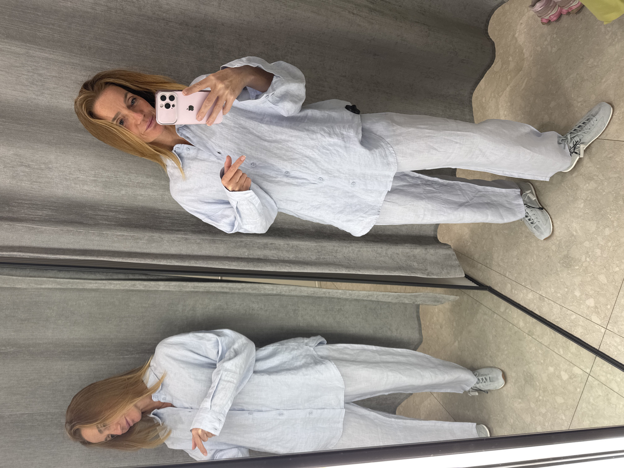 A woman with long, light brown hair is taking a mirror selfie, capturing her full body and reflection. She wears a relaxed-fit, light blue linen long-sleeved shirt with visible buttons and matching wide-leg pants. She is holding a pink smartphone, featuring the Apple logo on its back, to take the picture, while her other hand forms a small heart gesture. Her face, visible in the phone and her reflection, shows a subtle smile. On her feet are grey athletic sneakers with white soles and dark laces.
The setting appears to be an indoor space, likely a changing room or a retail environment. The background features grey textured curtains or wall coverings on one side, and a light-colored, possibly beige or off-white, tiled floor. A large mirror prominently displays her reflection, showing the outfit from a slightly different angle. Another pair of pink and beige sneakers is partially visible on the floor in the upper right background of the main image.