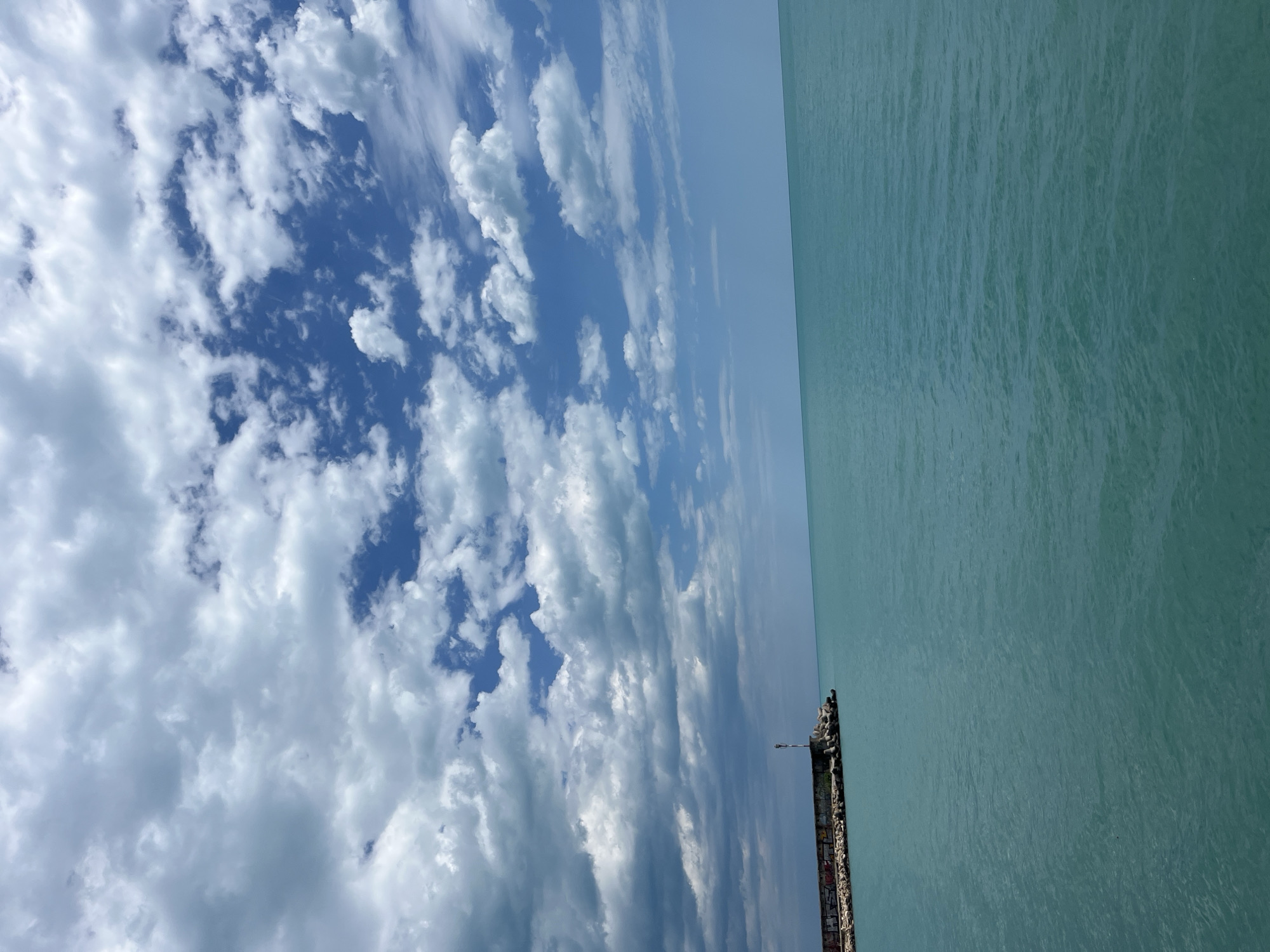 The image shows the Black Sea from the pier in Varna, Bulgaria. The sea occupies the right side of the image with a turquoise color and subtle wave patterns. On the left, a blue sky is partially obscured by white and gray cumulus clouds. A concrete pier or breakwater extends into the sea from the bottom center of the image. The pier is constructed of weathered concrete with visible graffiti. Several dark objects, potentially birds or structures, are positioned atop the pier near the water's edge.