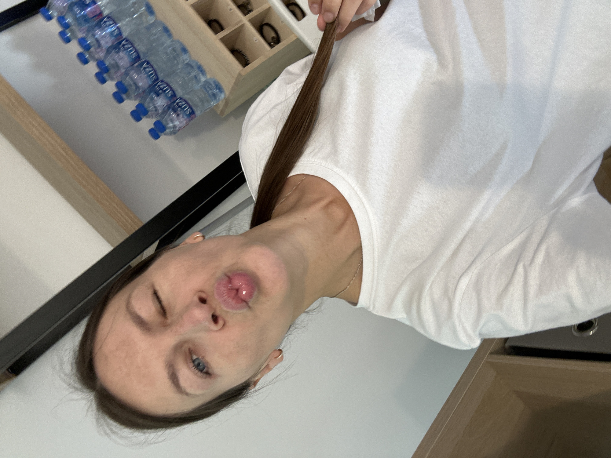 The image shows a young woman taking a selfie. She is winking and puckering her lips. She is wearing a white t-shirt and a silver necklace. Her hair is dark brown and pulled back. She is indoors, and in the background, there are several bottles of water on a shelf, as well as some small wooden compartments that appear to hold accessories. The overall impression is of a casual, personal moment. There is no indication of the location being Boreti, Montenegro.