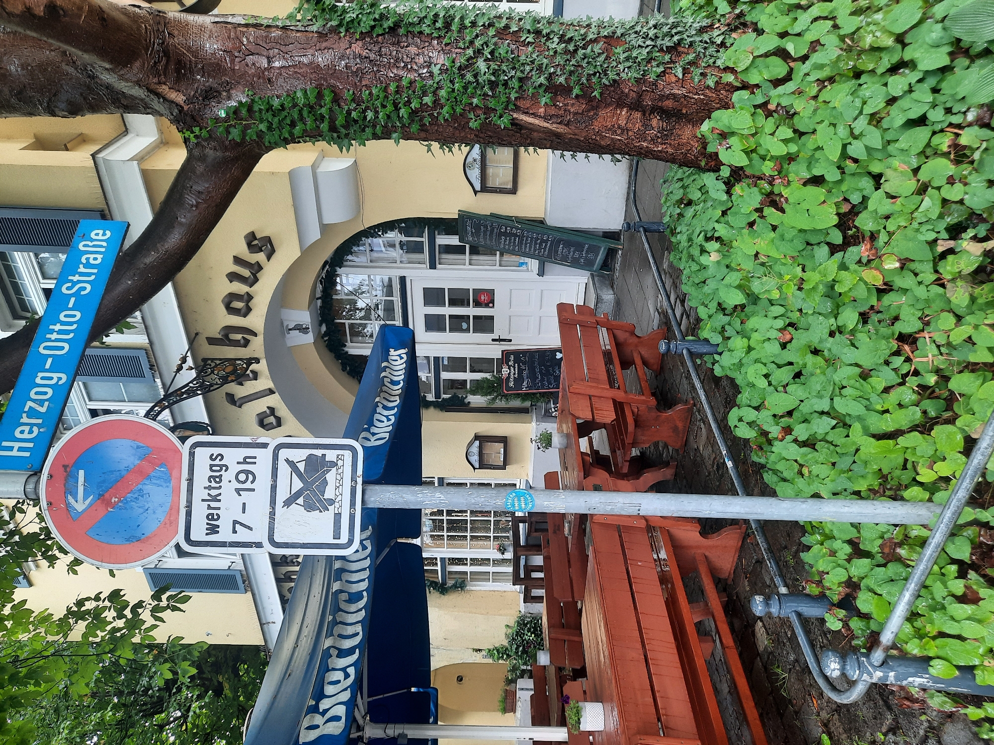 The image depicts an outdoor seating area of a restaurant or biergarten, likely in Rosenheim, Germany, as indicated by the street sign "Herzog-Otto-Straße". A blue awning with the word "Biergarten" is visible. Several wooden picnic tables and benches, stained red, are arranged on a paved area. In the foreground, a metal post holds traffic signs. The topmost sign is a circular red sign with a blue background and a white arrow pointing left, indicating no stopping or parking in that direction. Below it is a rectangular white sign with "werktags 7-19h" (weekdays 7 AM - 7 PM) and a crossed-out car symbol, signifying a restriction on vehicle access during those hours. Lush green ground cover and ivy climbing a tree are present. The building facade is yellow with arched windows and doors. A chalkboard menu stands near the entrance. The scene appears to be a public area, possibly a street-side cafe.