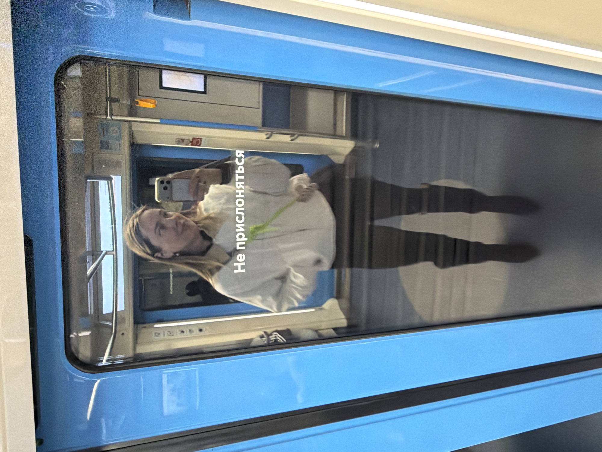 The image depicts the reflection of a person in the window of a metro train. The person is holding a smartphone and appears to be taking a photo or video of themselves. The train car's interior is visible through the window, revealing other seats and passengers, and the hallway in the adjacent car. A white inscription is visible on the window; “Не прислоняться” (Do not lean). The blue colour scheme of the train car is indicative of the Moscow Metro trains, specifically those used on the Bolshaya Koltsevaya line. The lighting suggests a daytime scene.