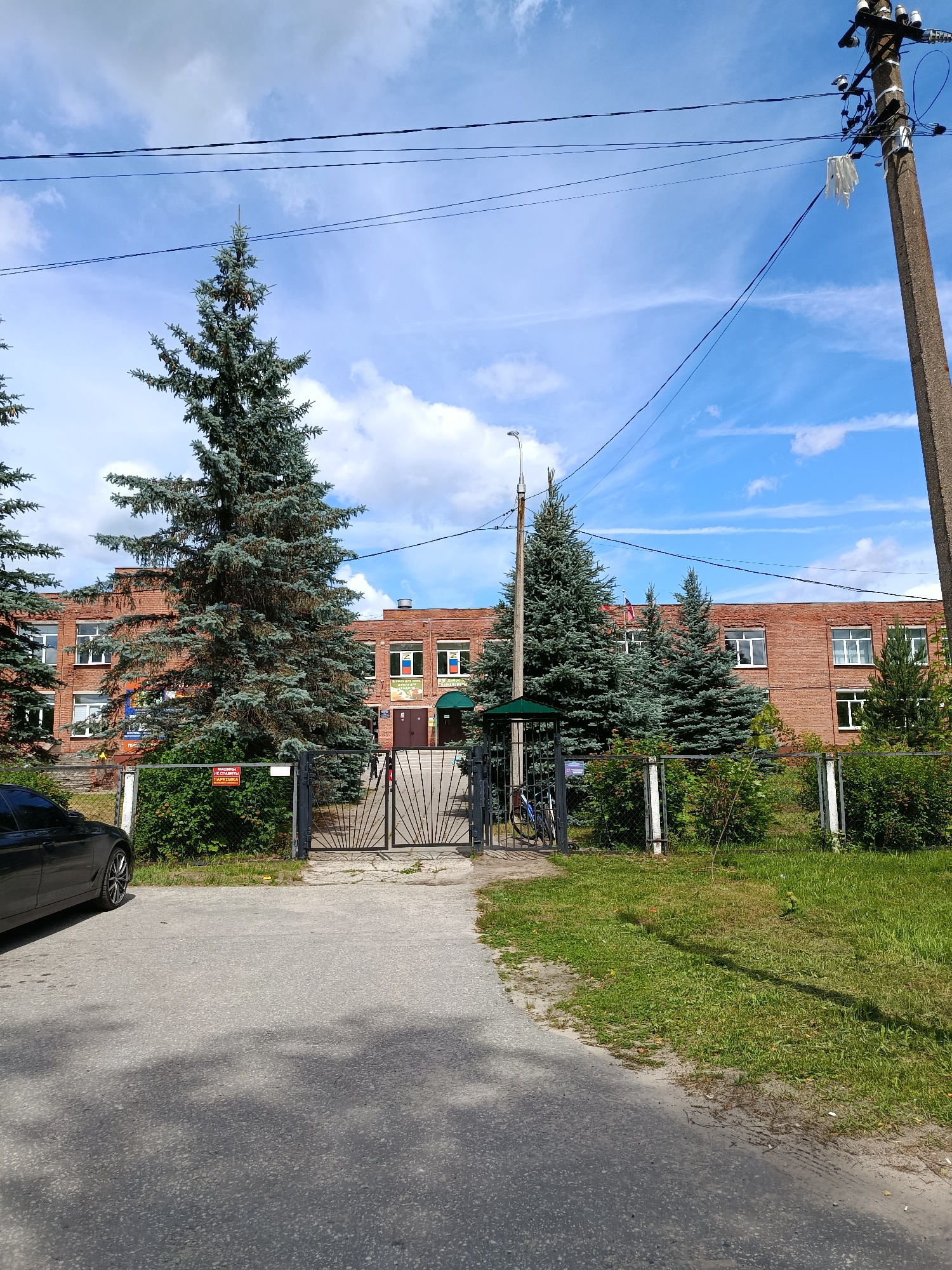The image depicts the entrance to a brick building in Vereya, Russia. Tall evergreen trees flank a metal gate leading to the building's entrance. A black car is parked on the left side of the frame. The building has multiple windows and appears to be a school or similar public institution, with flags and posters visible on its facade. The sky is blue with scattered clouds, and power lines run across the top of the image. A utility pole stands on the right side of the frame. There are no people actively engaged in any interaction within the frame. The scene is set on a sunny day, with shadows cast on the paved area in front of the gate.