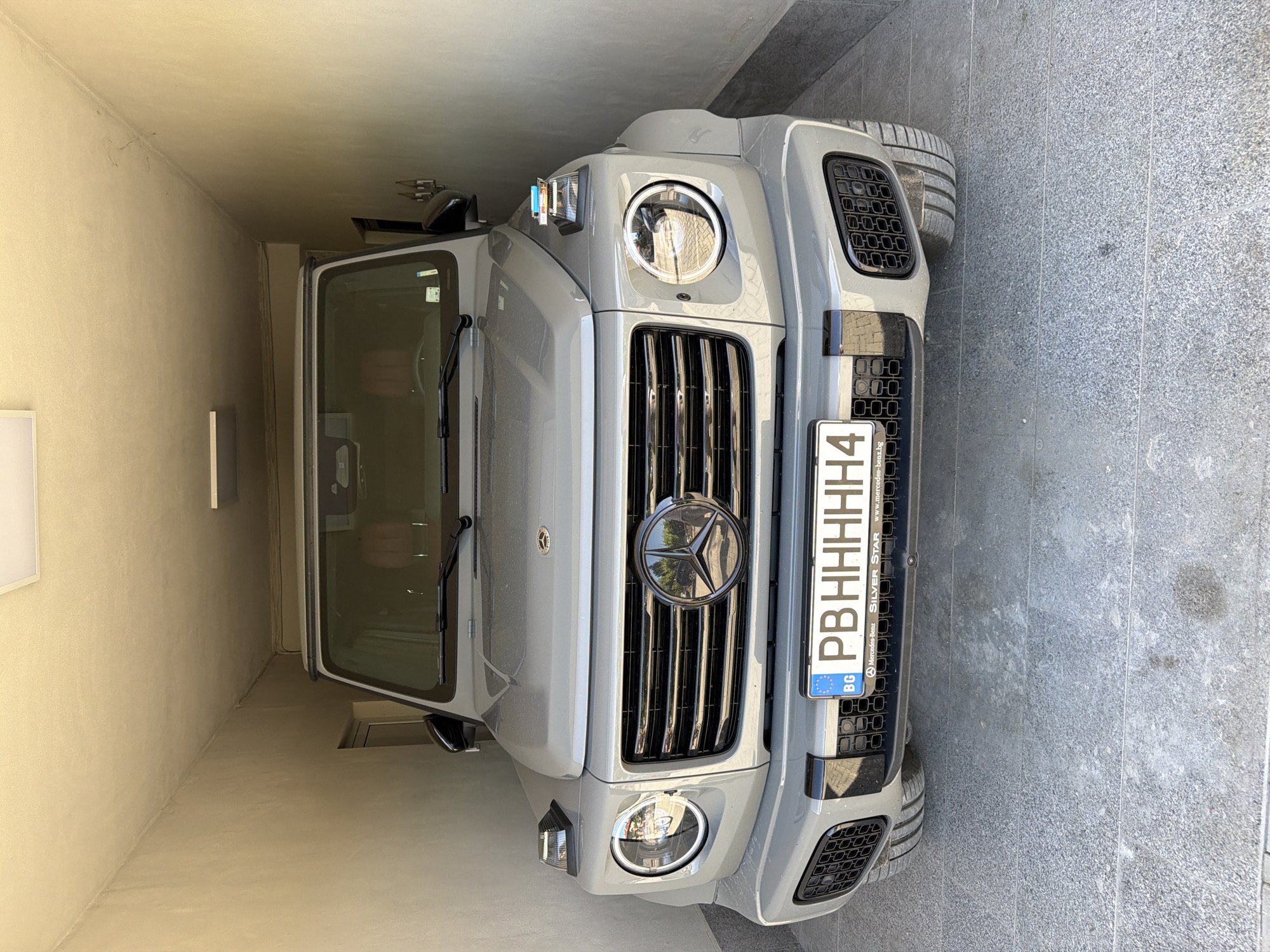 The image shows the front of a gray Mercedes-Benz G-Class SUV parked in a narrow space. The car's Bulgarian license plate reads "PBHHHHH4". The vehicle is located in an alley or tight parking spot with walls on either side. The location could be Asenovgrad, Bulgaria, as the license plate indicates a Bulgarian registration. The background is plain, with light-colored walls and a gray pavement. There are no visible people or other objects of significance.