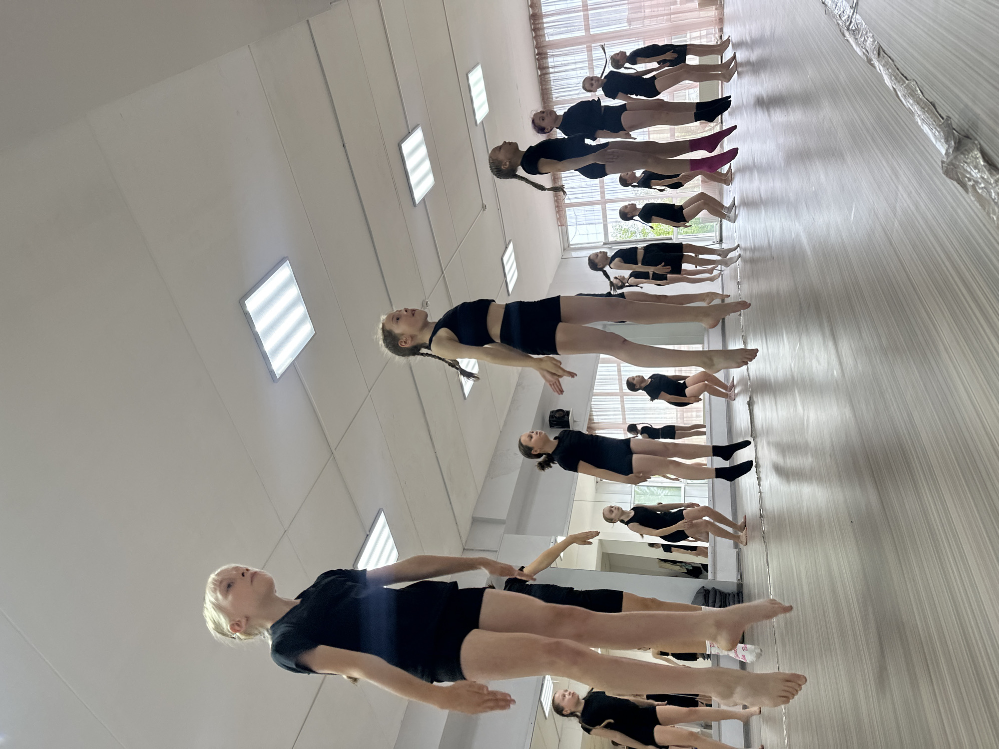 The media file depicts a group of young people, likely dancers, participating in a synchronized exercise or performance. They are dressed in black athletic wear, consisting of shorts and tops, and several individuals are wearing socks, one pair a striking pink. The setting appears to be an indoor studio or gymnasium, characterized by a pale, wood-paneled floor that reflects the subjects. The walls are white, with several rectangular fluorescent light fixtures illuminating the space. Large mirrors are visible along one wall, reflecting the group and creating a sense of depth and repetition. Through a large window or glass partition, greenery and possibly trees are visible, suggesting daylight. The subjects are positioned in a line, all performing a similar pose where they are levitating or balancing with their bodies parallel to the floor, their arms extended or held in specific positions. Their faces show concentration and focus, with some looking upwards. The overall atmosphere seems disciplined and professional, indicative of a dance class or rehearsal. There is no visible text in the image. The lighting suggests it is daytime, and the indoors setting implies the weather is not a factor.