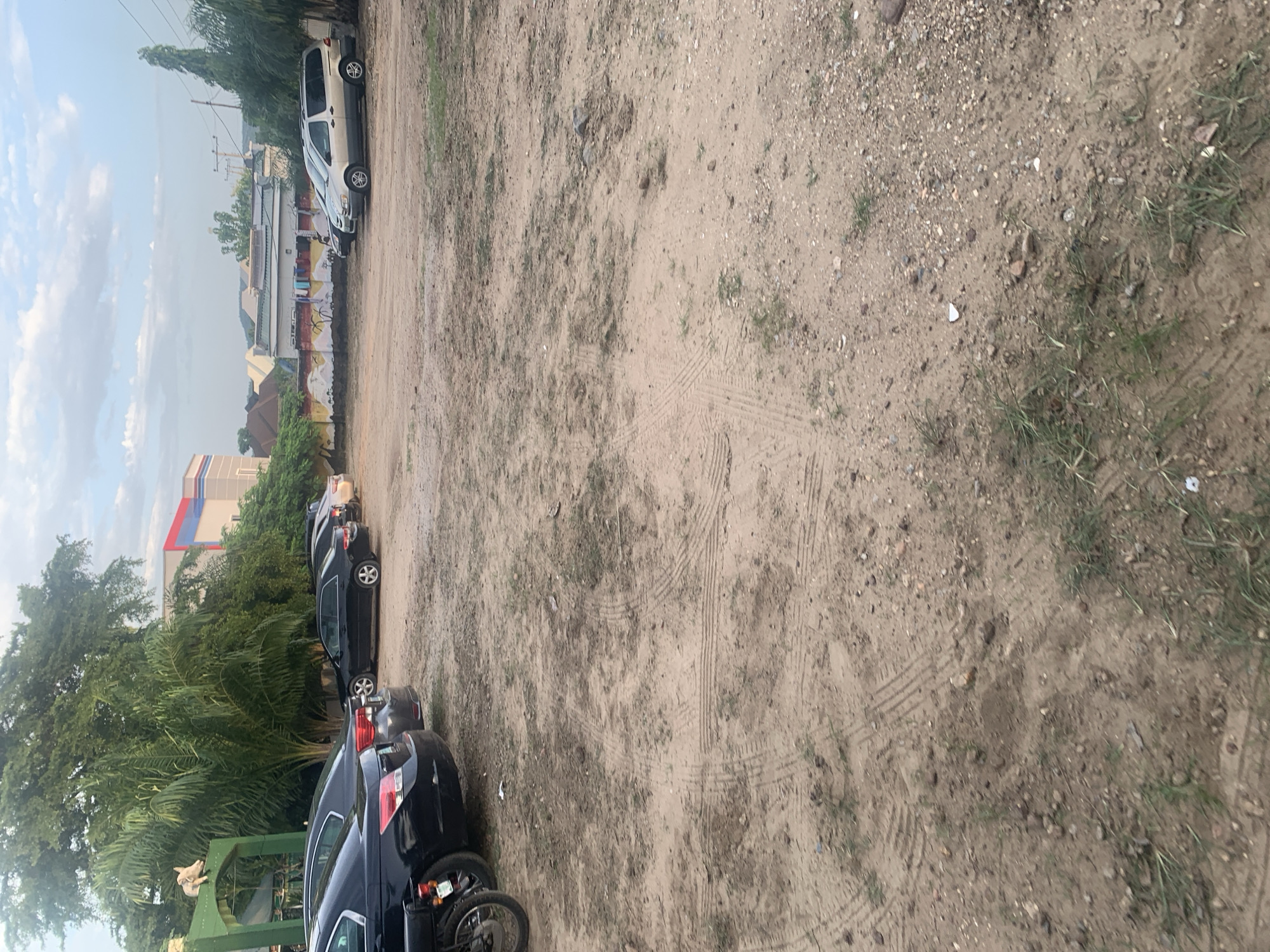 The image shows an outdoor parking lot in Adankolo, Nigeria. The ground is a mixture of dirt and gravel, with visible tire tracks from vehicles. Several cars are parked in the lot, including black sedans and a light-colored minivan. In the background, there are buildings, trees, and power lines. The sky is partly cloudy. There are no people visible in the image.