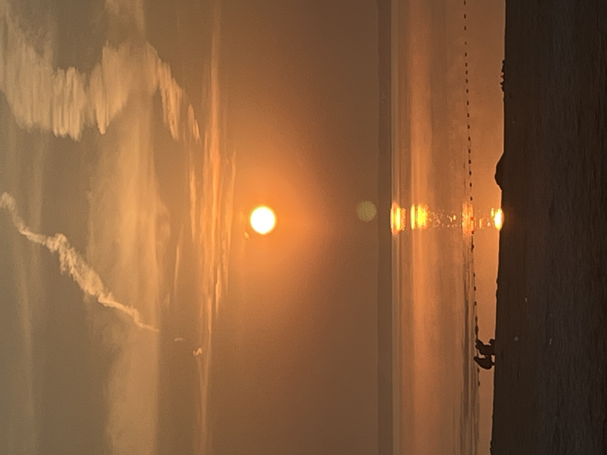 The image depicts a sunset over a body of water, possibly a sea or a large lake, near the city of Surakovo, Russia. The sun, a bright orange orb, is positioned slightly above the horizon, casting a warm glow across the sky. The sky is filled with a mix of orange and yellow hues, with white contrails from an aircraft creating abstract patterns. In the foreground, a dark silhouette of a sandy shore is visible. Two figures, also in silhouette, are crouched on the shore, facing the water. The water reflects the vibrant colors of the sunset, creating a shimmering effect. A dotted line, possibly a marker or a boundary, is faintly visible across the water. The overall scene conveys a sense of calm and serenity.