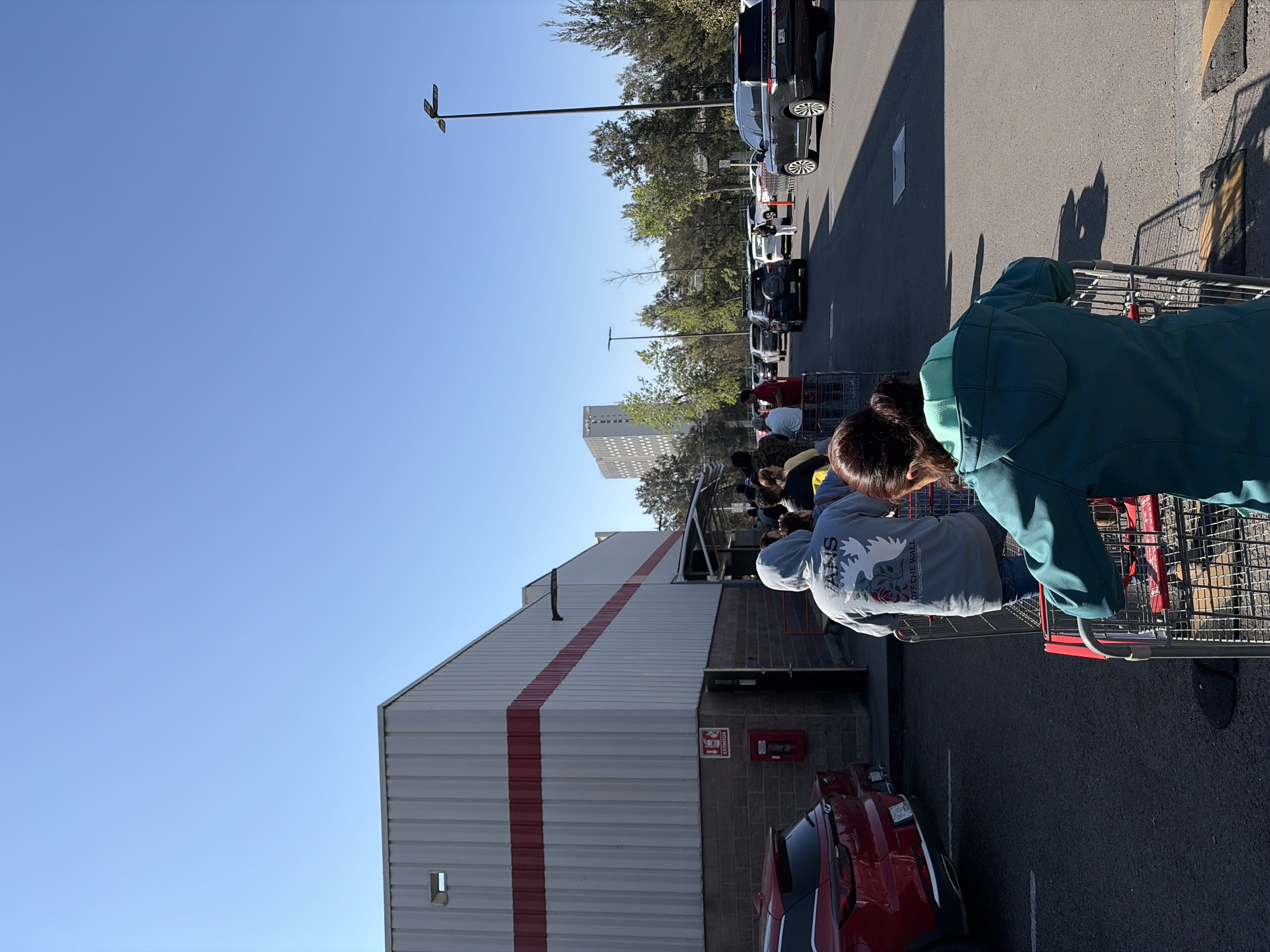 The image shows a long queue of people with shopping carts lined up outside a building. The building is large with a white exterior and a thick red stripe along the side. A red car is parked near the building, and behind the building, a tall apartment complex is visible. Cars are parked along the street. The sky is blue, and the scene appears to be taking place in an urban environment. Based on the fire extinguisher graphic, it is probable the location is Mexico, and the presence of Vans and "off the wall" apparel suggests the location is outside the USA. Given the architectural style of the apartment complex and the retail building, it is probable the location is the city of Puebla, Mexico. People are standing and waiting in line, likely to enter the building. They are interacting by standing in a line, but there are no other obvious interactions.