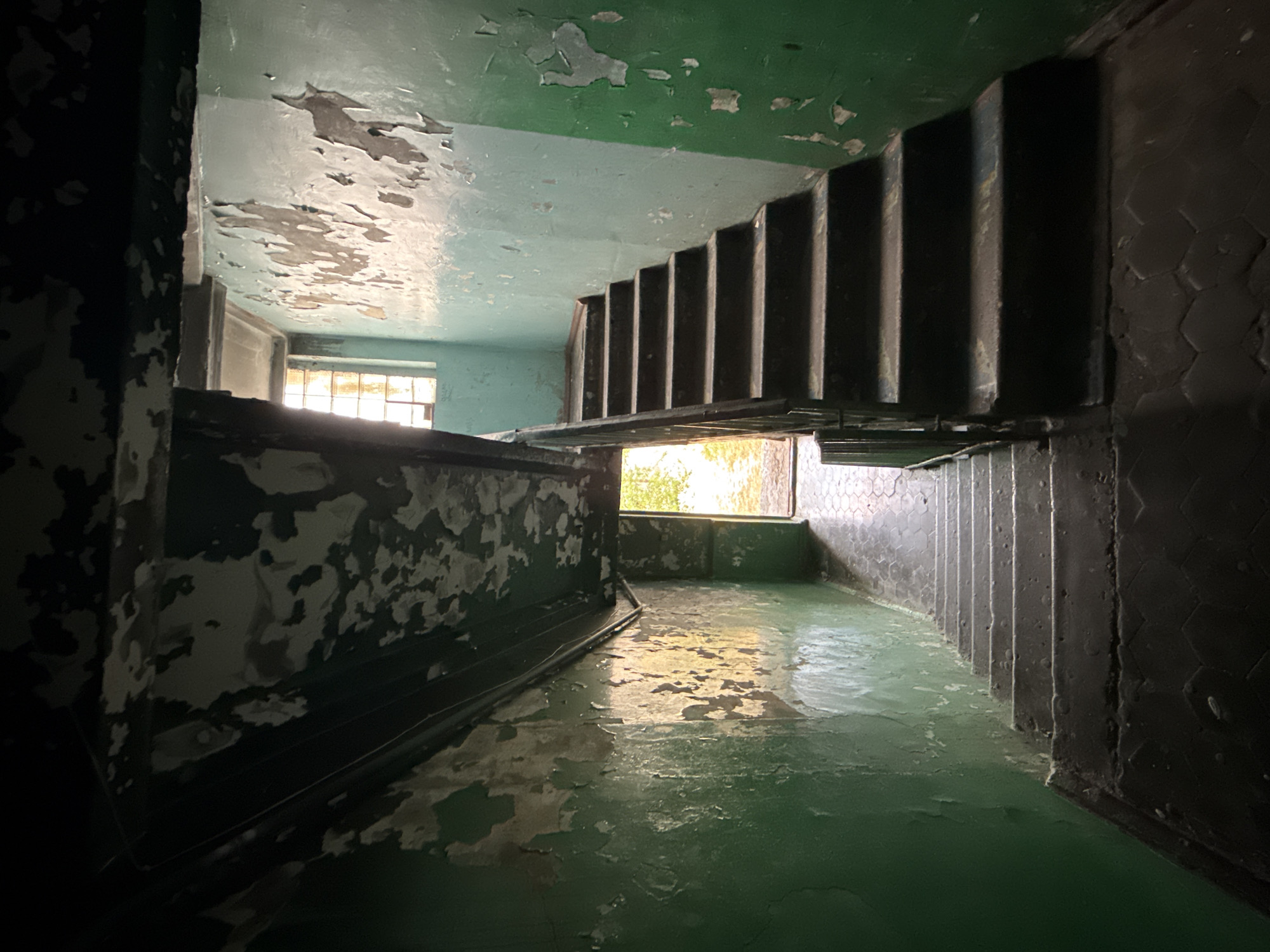 This is a photograph taken inside an abandoned stairwell, likely in Novorossiysk, Russia. The scene is dimly lit and shows signs of decay and neglect. The walls are painted a peeling green, with patches of plaster exposed. There are two staircases visible; one leading upwards to the right and another leading downwards to the left. The stairs are dark and appear worn. At the top of the right staircase, there is a window with a grid pattern and natural light coming through. An open doorway is visible at the end of the left staircase, through which faint greenery and sunlight can be seen, suggesting an exit to the outside. The floor is tiled with a hexagonal pattern. There are no people or animals present in the image. The overall impression is one of disuse and abandonment.