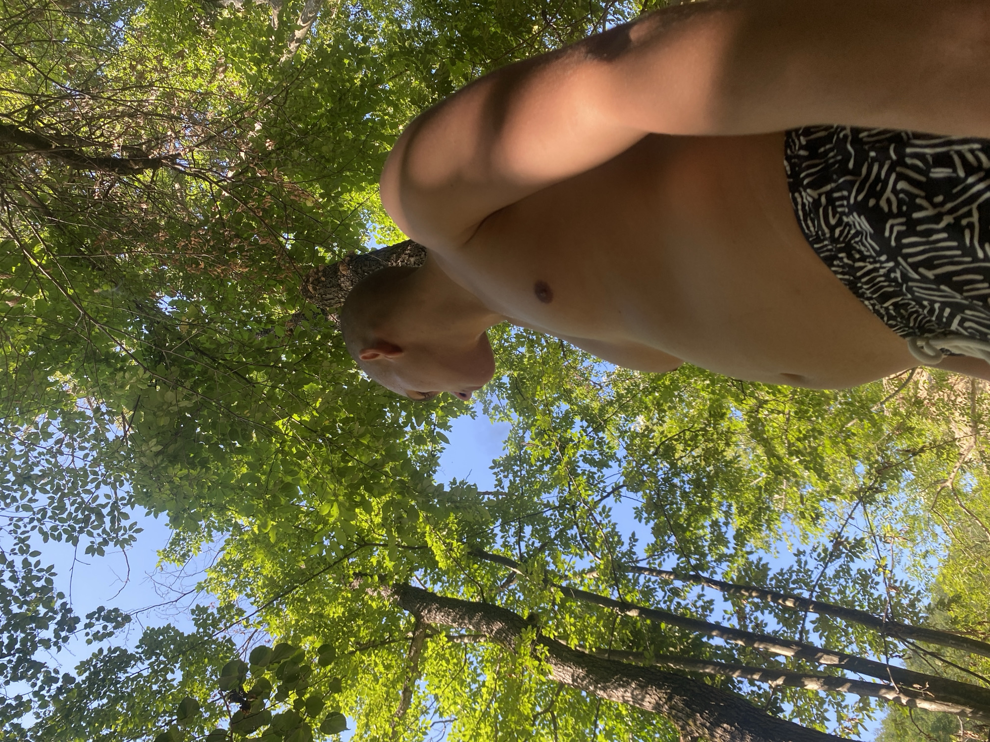 A young person is shown from a low angle, looking up at the sky through a canopy of green trees. The person is shirtless and wearing black and white patterned shorts. Sunlight filters through the leaves, creating dappled shadows on the person's skin. The sky is a clear blue. The location is not specified as Debnevo, Bulgaria.