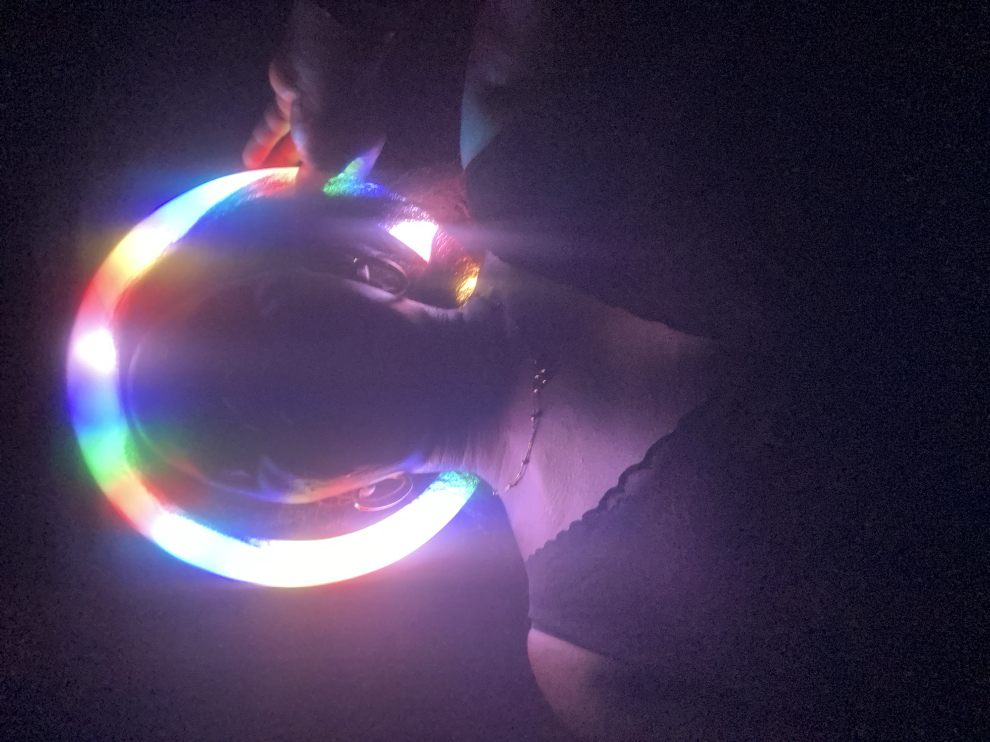 A person is holding a multicolored light ring near their face in a dimly lit or dark environment. Part of their upper body, including the neck and shoulder, is visible. A necklace adorns the neck. The environment appears dark, with only the light ring illuminating the immediate surroundings. The location is not specified, making it impossible to ascertain if it is in Maigizo, Nigeria.
