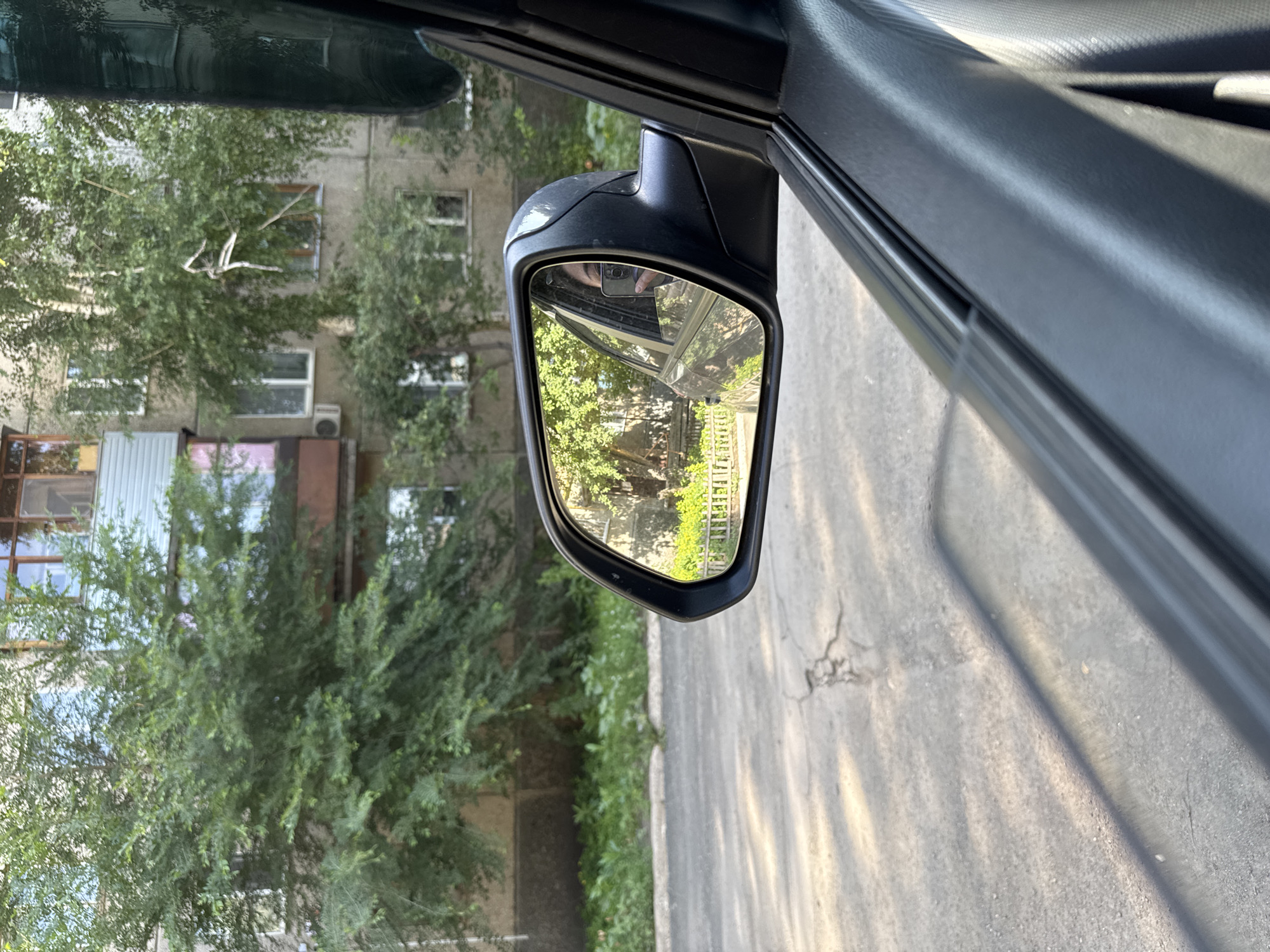 An exterior shot from inside a vehicle shows a side-view mirror reflecting trees and a building. The mirror is black with a silver trim. The building is light gray with multiple windows and balconies. Lush green trees are in front of the building, with sunlight filtering through the leaves. The road in the foreground is asphalt with some cracks. The image is taken from the perspective of a person sitting in the driver's or passenger's seat of a car. The car's interior, including a black door panel and a portion of the window frame, is visible in the foreground. The scene appears to be in a residential area of Novotroitsk, Russia, likely during daylight hours given the bright sunlight and shadows.
