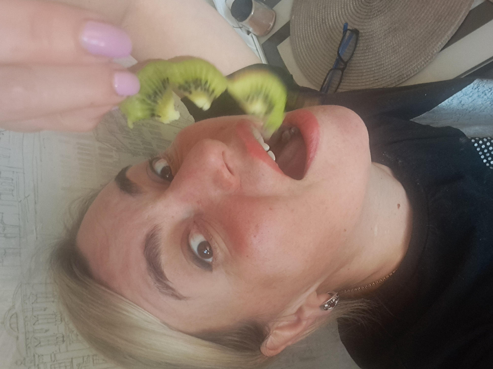 A woman is lying on a surface with a patterned covering, possibly a bed or table. She is in the process of eating kiwi fruit, which is being held in front of her open mouth by a hand with light pink nail polish. The woman has fair skin, blonde hair, and is wearing an earring and a necklace. She is wearing a black top. In the background, there is a metallic container, a round woven mat, and a pair of glasses. The location appears to be indoors, possibly a home setting. There is no information to suggest the image was taken in Elektrostal, Russia.