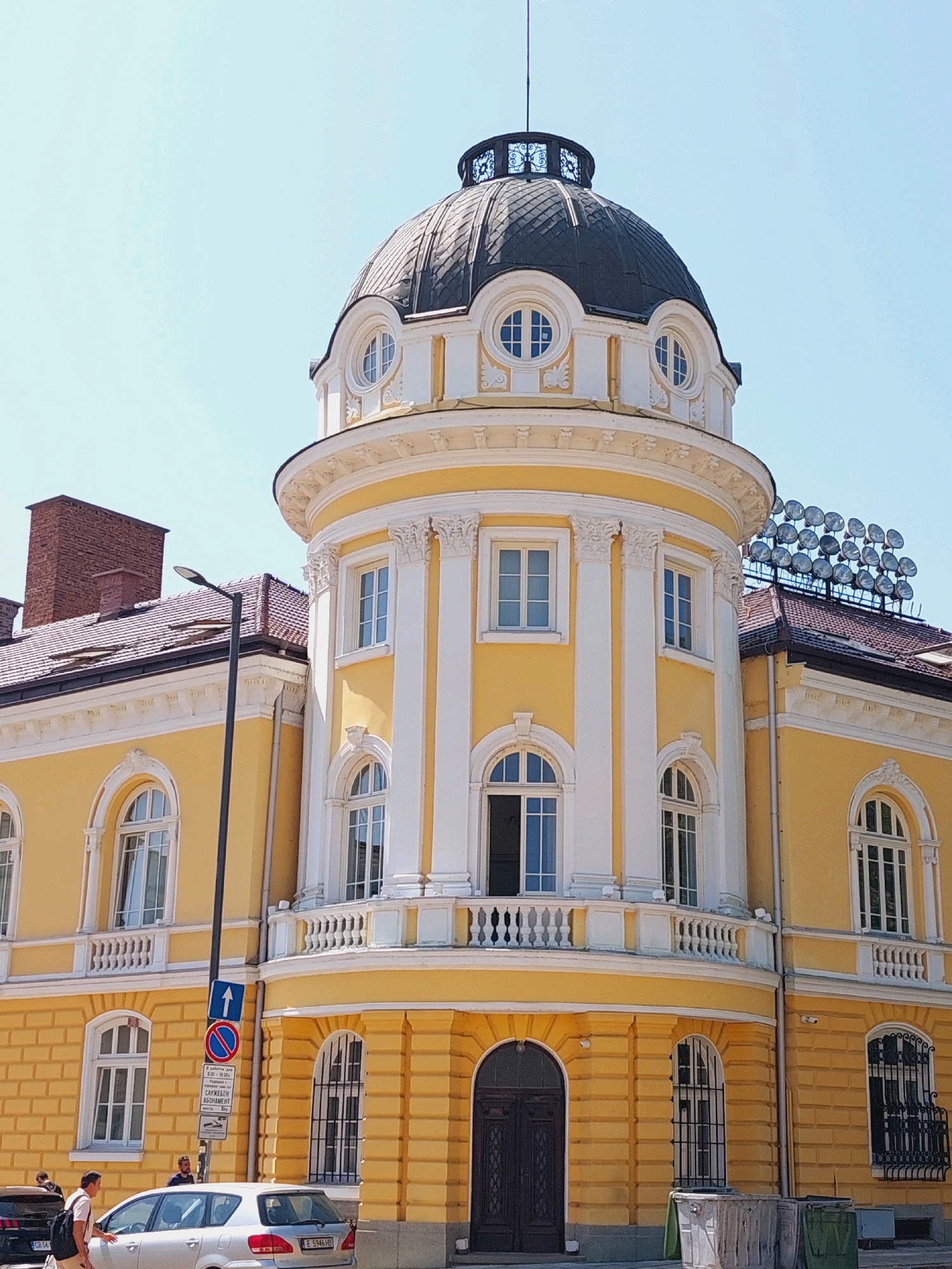 The image shows the main entrance and a corner section of the National Theatre "Ivan Vazov" in Sofia, Bulgaria. The building is predominantly yellow with white decorative elements, columns, and a black dome. Several windows, some with arched tops and ornate grills, are visible. A white balcony with decorative railing spans across the curved section of the building. In the foreground, a silver car with a Bulgarian license plate is parked, and two individuals are walking past it. Further down the street, another car is parked. A street lamp and traffic signs are positioned next to the building. The sky is clear and blue.