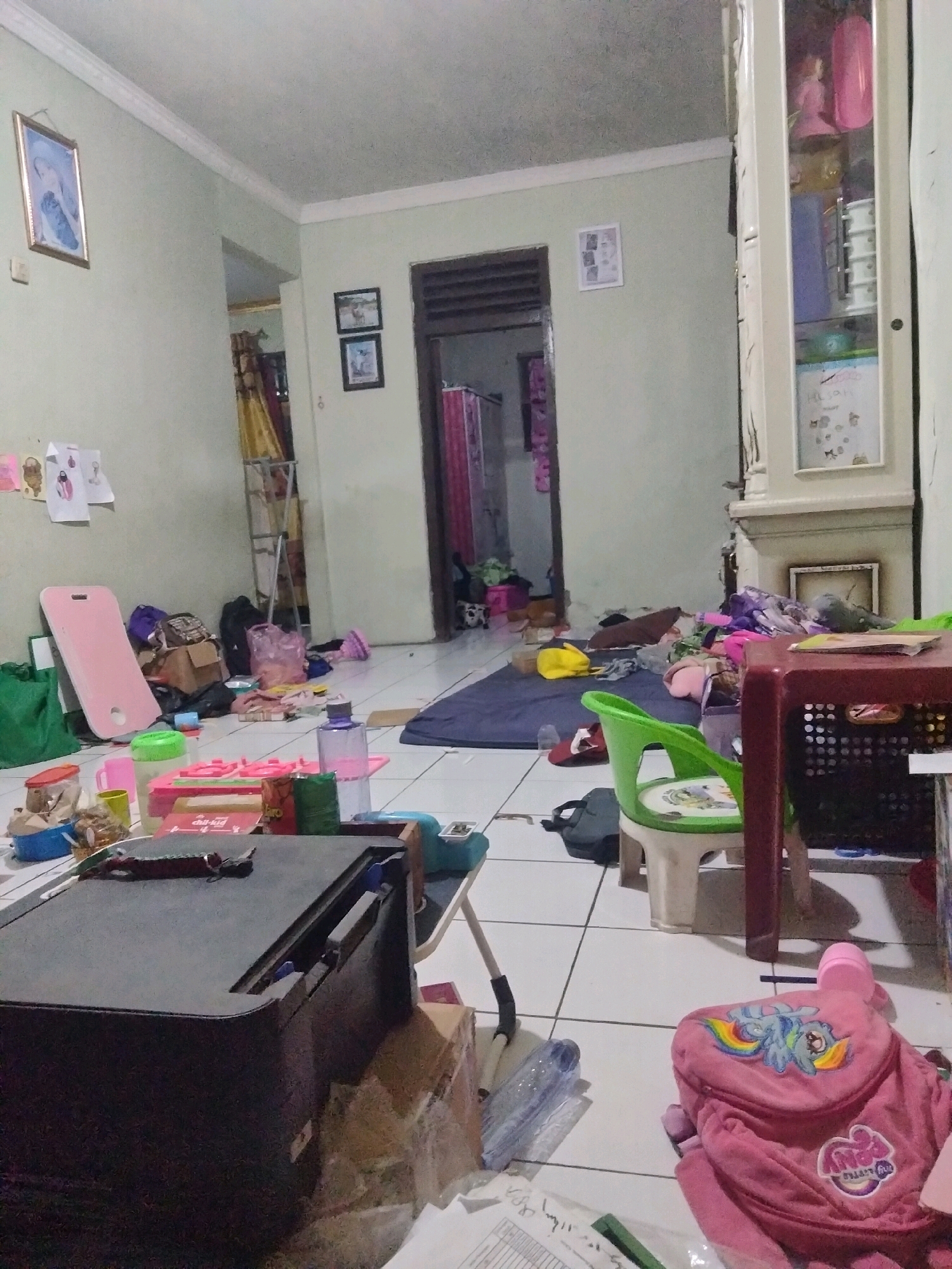 The image depicts a cluttered room, likely a child's play area, in a residential setting in Suka Sari, Indonesia. The floor is tiled and strewn with various objects, including toys, bags, boxes, and what appears to be a printer. A pink play mat leans against a wall, and a small green plastic chair with a child's drawing on the seat is prominent. In the background, an open doorway reveals a glimpse of another room with curtains. A tall, narrow cabinet with glass doors stands to the right, displaying colorful items within. A pink backpack with a "My Little Pony" design is visible in the foreground on the right. The overall impression is one of disarray and active use, suggesting a domestic environment where children's activities take place.