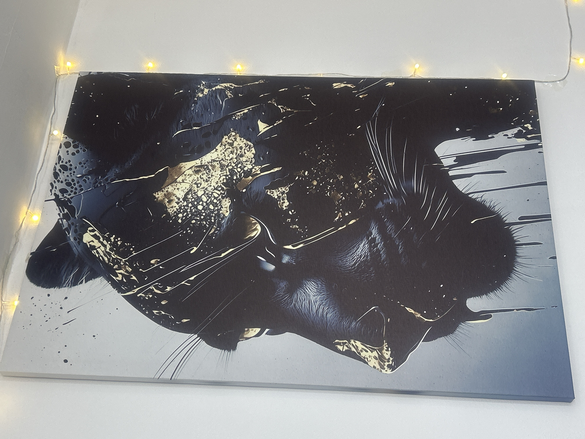 The image shows a canvas print of a black panther's head, rendered in a stylized manner with black and gold paint splatters and drips. The panther is depicted in profile, facing left. The background of the print is a gradient of light grey to blue. The canvas is hanging on a white wall, and a string of small, yellow fairy lights is draped along the top edge of the wall, partially visible in the upper right corner of the image. The image does not contain any information about the city of Almaty, Kazakhstan, or any specific location.