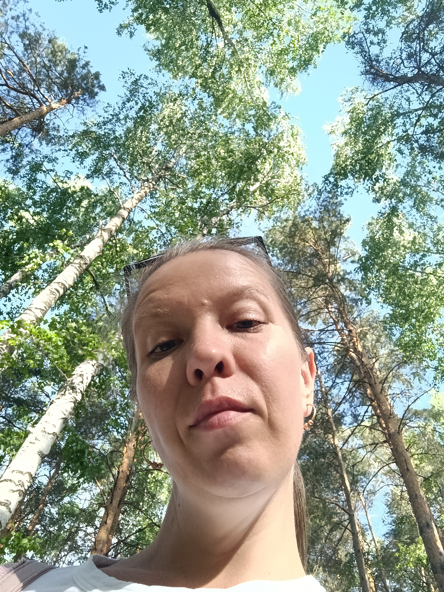 A selfie was taken of a woman from a low angle, in an area with birch and pine trees. The sky is visible through the canopy of leaves. She is wearing earrings. Her glasses are propped on top of her head. The location may be in Chaikovsky, Russia, given the prompt.