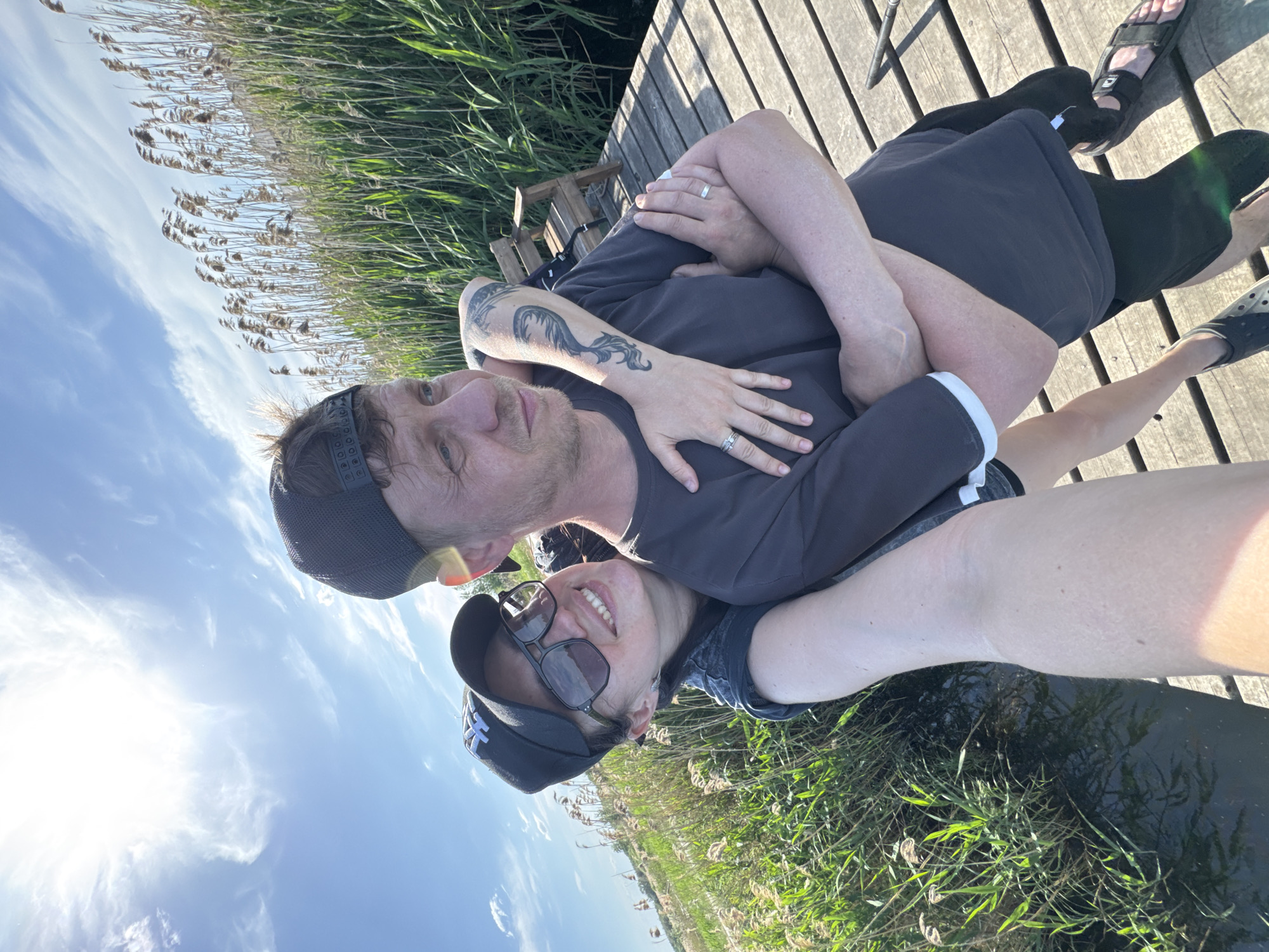 A couple is on a wooden boardwalk near tall grass. The man has tattoos on his arm and both people are wearing rings. The woman is wearing sunglasses and a baseball cap. The environment includes water, sky, and plants. The city is Unknown, Unknown.