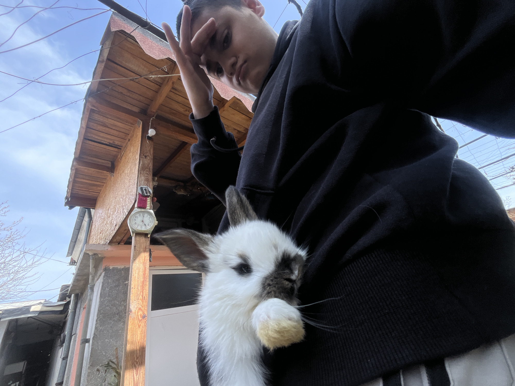 A low-angle selfie shows a person holding a white rabbit in front of them. The person has dark hair and is wearing a dark hooded sweatshirt. The rabbit is mostly white with some darker markings around its eyes and ears. The rabbit is holding its paw up to its mouth. In the background, a building with a wooden roof and siding is visible, along with a watch hanging from a post. The sky is blue with some clouds. The location is Lozarevo, Bulgaria.