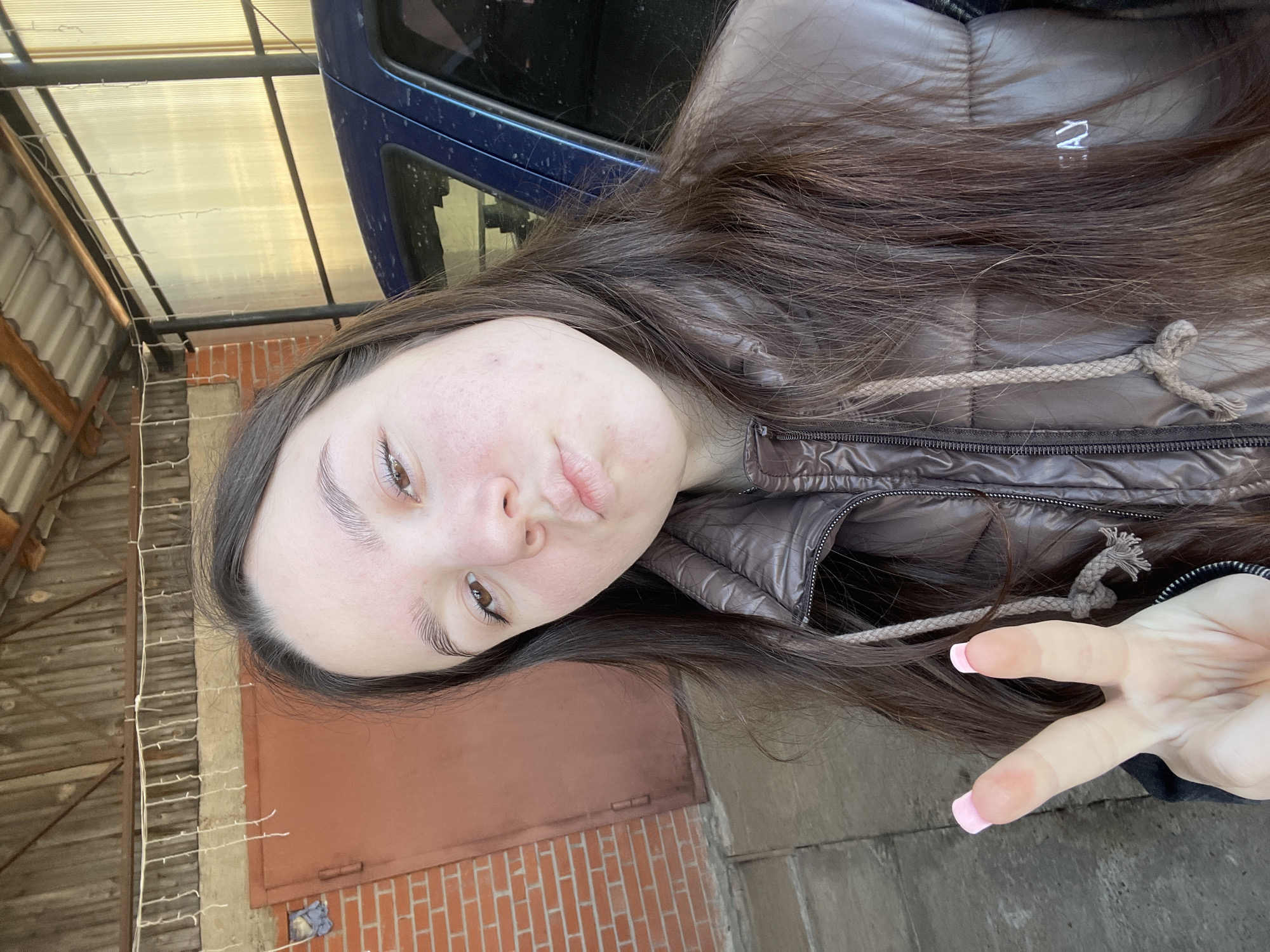 A female subject is captured in a front-facing, waist-up portrait in what appears to be a sheltered outdoor area in the city of Фоки, Russia. The individual has long, dark brown hair, brown eyes, and a light complexion with visible facial blemishes. She is wearing a dark brown, possibly metallic-sheen, puffer jacket with a visible zipper and a drawstring with a knot near her left shoulder. Her lips are pursed forward. Her left hand is raised to her chest height, displaying two fingers in a "V" or "peace sign" gesture, with pink painted fingernails on the index and middle fingers.
The background behind the subject reveals an environment consistent with a private residence or a workshop area. To the left of the subject, a reddish-brown, possibly metal, double door is visible, set into a wall constructed of red bricks. Above this, a string of white, icicle-style decorative lights is strung horizontally against a lighter colored wall section, beneath a wooden roof structure with horizontal planks. To the right of the subject, a dark blue vehicle, likely a car, is partially visible, appearing to be parked under a transparent or translucent corrugated plastic roof panel, supported by dark metal posts. The ground visible at the bottom left appears to be a concrete surface.