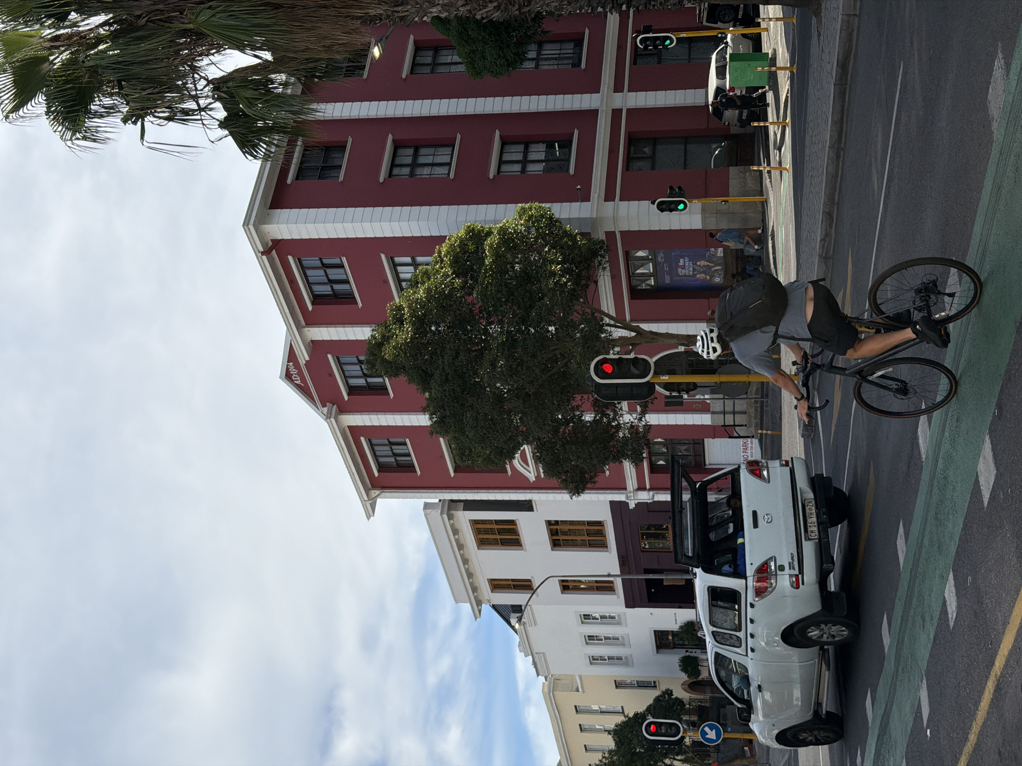 A cyclist rides a bicycle on a green painted bike lane along a city street in Cape Town, South Africa. The cyclist wears a helmet and a backpack. A white Mazda pickup truck with open bed is parked on the street near a traffic signal. Adjacent buildings have red, white, and beige facades. Traffic lights are visible, some displaying red and others displaying green. The sky is overcast. A palm tree is visible in the upper left corner.
