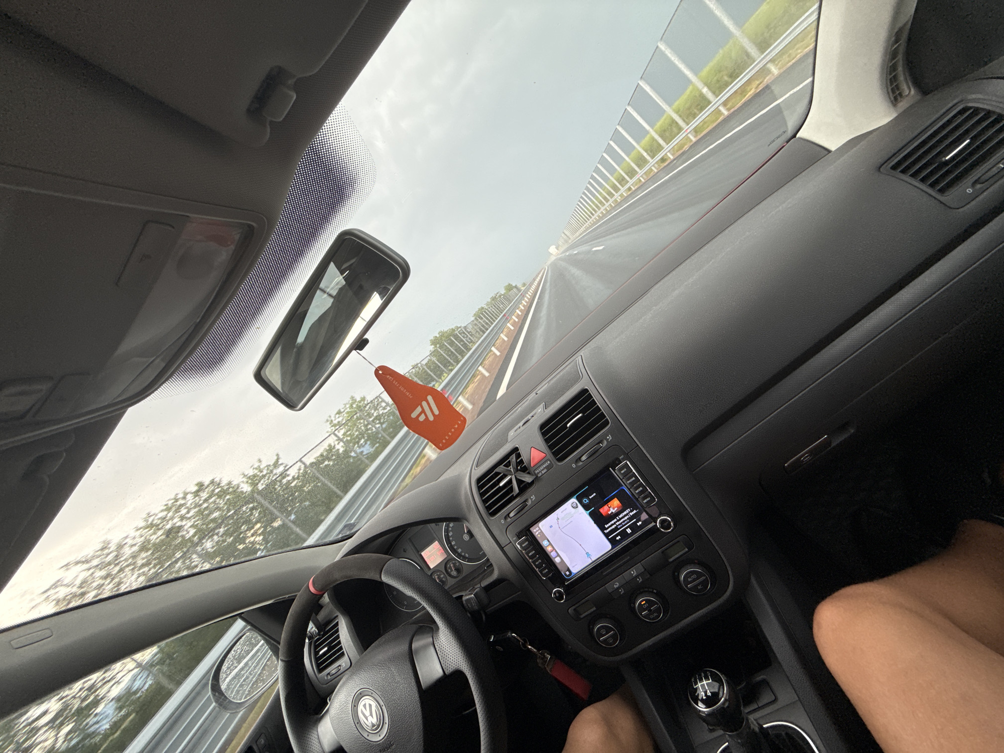The image provides an interior view of a car, likely a Volkswagen Golf, from the passenger's seat while driving on a highway near Chokoba, Bulgaria. The perspective is tilted, looking forward and slightly right.
The scene outside the car shows a multi-lane highway stretching into the distance, flanked by a sound barrier on the right and trees beyond. The sky is overcast and appears bright, with some water droplets on the windshield suggesting recent rain or damp conditions.
Inside, the car's dashboard, steering wheel, and central console are clearly visible. The steering wheel features the Volkswagen logo and has a section with red stitching or wrapping. An orange, leaf-shaped air freshener, with the brand name "FRESHWAL" or similar starting with "FREE," hangs from the rearview mirror. A manual 6-speed gear shifter is prominent in the lower center. The infotainment screen displays a navigation map on the left and a music player on the right. The music player shows "Тупак Шакур - Калифорния Лав" (Tupac Shakur - California Love) playing, indicating Cyrillic script. Other visible text on the screen includes "Q Search" and buttons for "PHONE," "RADIO," "MEDIA," "MENU," and "SOUND." Climate control knobs are marked "AUTO," "AC," and "DUAL." A "PASSENGER AIR BAG OFF" indicator is also present. Parts of a person's legs or knees are visible in the foreground, suggesting at least two occupants in the vehicle.
The image captures a typical road trip or commute, emphasizing the car's interior details and the open road.