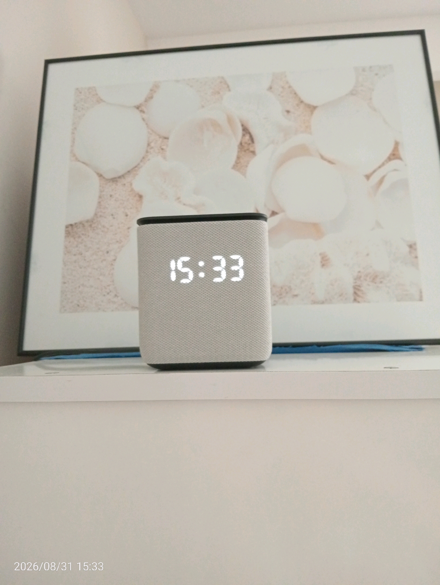 The image shows a digital clock displaying 15:33, placed on a white shelf. Behind the clock is a framed picture of what appears to be light-colored shells on a sandy background. The overall scene is indoors, with a neutral-toned wall in the background. There is a date stamp at the bottom left of the image that reads "2026/08/31 15:33." There is no information in the image to suggest a location in Silifke, Turkey.