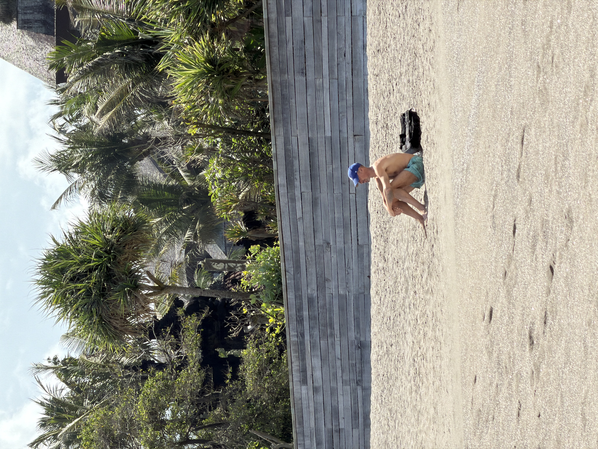 A man wearing a blue baseball cap and teal swim trunks sits on a sandy beach in North Kuta, Indonesia. He is facing left, with his arms resting on his knees and his legs bent. Next to him, on his right, is a black duffel bag. Behind the man is a wooden retaining wall made of horizontal planks. Beyond the wall, there is lush tropical vegetation including palm trees and other dense foliage. The sky above is partly cloudy. The beach sand in the foreground is light brown and shows scattered footprints.