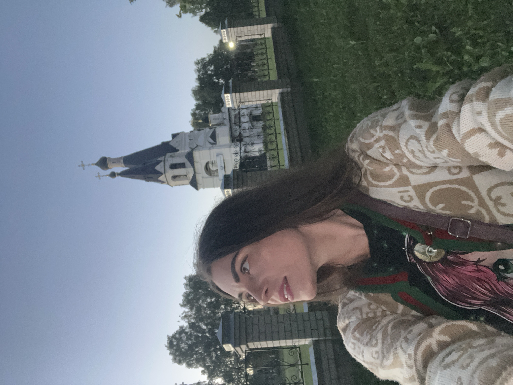 This is a vertically oriented, low-angle, close-up selfie of a young woman taken outdoors. She is in the foreground, her face and upper body are visible. She has long, dark hair and is looking to her left, with a slight smile. She is wearing a patterned sweater over a shirt with a colorful graphic print and a dark strap across her chest.
In the background, to her left and slightly behind her, is a white building with dark roofs and crosses on top, resembling a church or a monastery. A black metal fence with decorative elements separates the building from a patch of green grass and trees. The sky is a clear, pale blue, indicating it is daytime, likely late afternoon or early evening due to the soft light. The overall scene appears peaceful and serene. The image appears to be taken in Krasnoe-na-Volge, Russia. There is no visible text in the image.