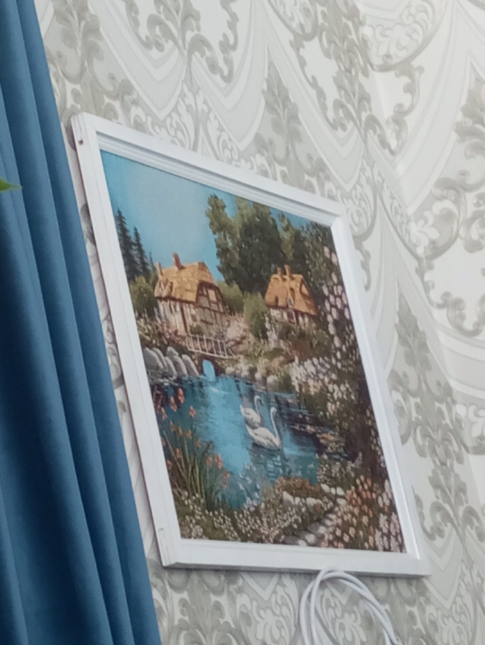 The scene, stated to be from Yaromaska, Russia, depicts an interior wall featuring a framed picture and elements of room decor. The image itself provides no visual information to corroborate its stated geographical location.
The primary subject is a rectangular picture in a white frame, mounted on a wall covered with light-toned wallpaper featuring a repeating grey ornamental or scroll pattern. The picture, which appears to be a painting or print, depicts a pastoral landscape scene. Within the artwork, two thatched-roof cottages are visible, one situated on the left and another on the right, set amongst numerous green trees. A small wooden bridge spans a calm body of blue water, where two white swans are seen swimming in the lower-middle section. The banks of the water and the foreground are densely populated with various flowering plants and rocks.
To the left of the framed picture, a segment of a vertical, pleated blue curtain is visible. Below the lower right corner of the picture frame, a coiled white electrical cable is present against the wallpaper. The photograph is captured from an oblique angle, looking slightly upwards and to the right, causing the framed picture to appear slightly skewed from a frontal perspective.