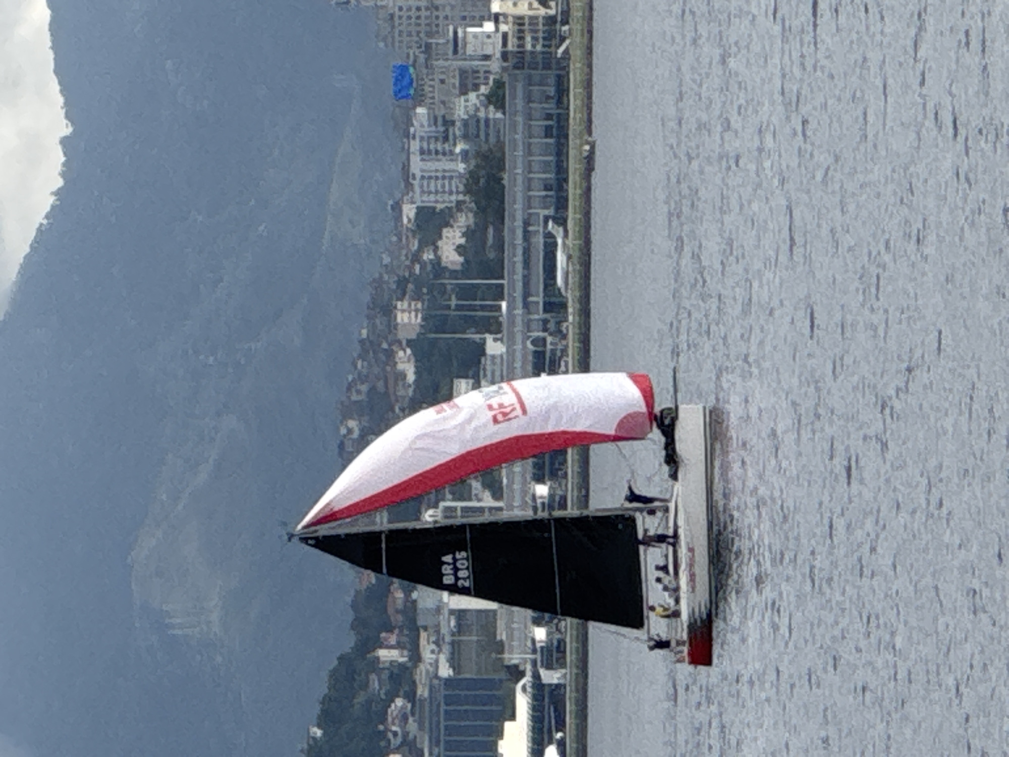 The image captures a sailboat with black and white sails, featuring red accents, navigating on the water. The sail displays the text "BRA 2805". A group of people are visible on the deck of the sailboat. In the background, a city skyline with buildings and hills can be observed, suggesting an urban coastal environment. The presence of buildings and hills in the background, along with the specific sailboat markings, points to a location in Niterói, Brazil, likely within Guanabara Bay, a common area for sailing activities and offering views of Rio de Janeiro's distinct topography. The overall scene depicts a daytime maritime activity against a backdrop of urban development and natural landscape.