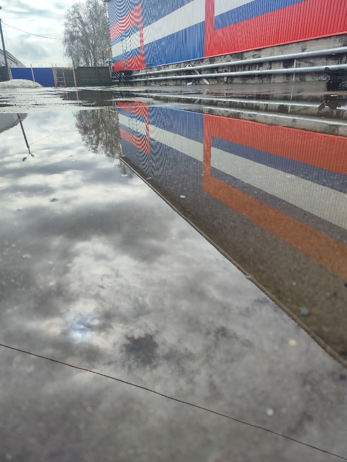 An outdoor scene in Chaikovsky, Russia, is captured from a low perspective, with a large puddle dominating the foreground. The puddle’s surface acts as a mirror, reflecting the cloudy, overcast sky and a prominent building facade. Patches of brighter light within the reflected clouds suggest the sun attempting to break through. A thin, dark line, possibly a power line or cable, stretches horizontally across the lower portion of the reflected sky.
Above the puddle, the wet ground, likely asphalt or concrete, extends towards the midground where a building stands. The building's exterior features corrugated metal siding painted in distinct horizontal stripes of red, white, and blue, strikingly resembling the Russian national flag. The red stripe is at the top, white in the middle, and blue at the bottom. Below the striped wall, multiple horizontal pipes or conduits are visible.
In the background, to the left of the building, bare trees with intricate branches stand against the cloudy sky. Fences and other undefined structures can be seen further in the distance, suggesting an urban or industrial setting. The wet ground and the leafless trees indicate a cool season, possibly late autumn, winter, or early spring. The time of day is daylight, under a predominantly overcast sky. There are no people or discernible text in the image.