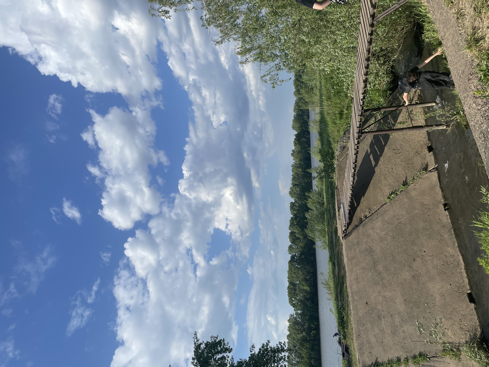 The image presents a landscape under a partly cloudy, blue sky. A body of water, likely a river or wide stream, extends into the distance, bordered by a line of trees. A concrete structure, perhaps a retaining wall or part of a bridge foundation, is present in the foreground, with some vegetation growing on and around it. A wooden structure is also partially visible. A person is present near the water. The location is unspecified, and there are no clues to determine the city or area.