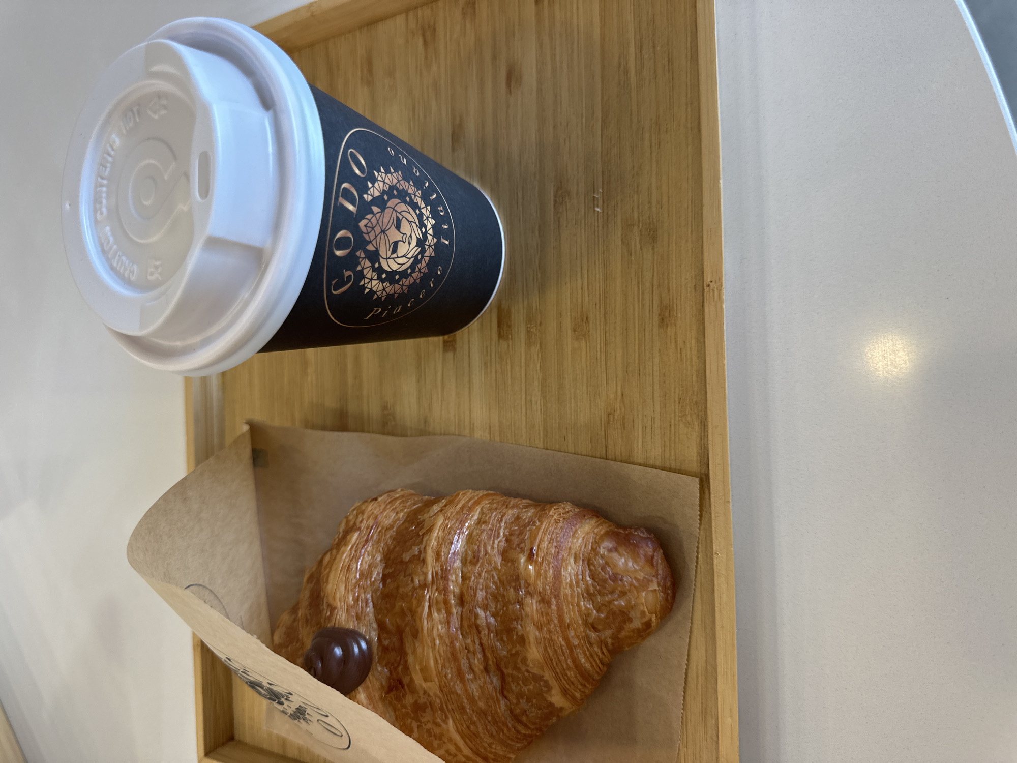 A high-angle close-up shot shows a dark brown disposable coffee cup with a white lid and a flaky croissant on a light brown bamboo tray. The cup has a gold logo with the text "GODO Piacere Italiano" and an image of a woman's face. A small dollop of dark brown chocolate cream is visible on top of the croissant. The tray is placed on a white speckled countertop, likely in a cafe or restaurant in Rafailovići, Montenegro. The background is a blurred white wall and ceiling.