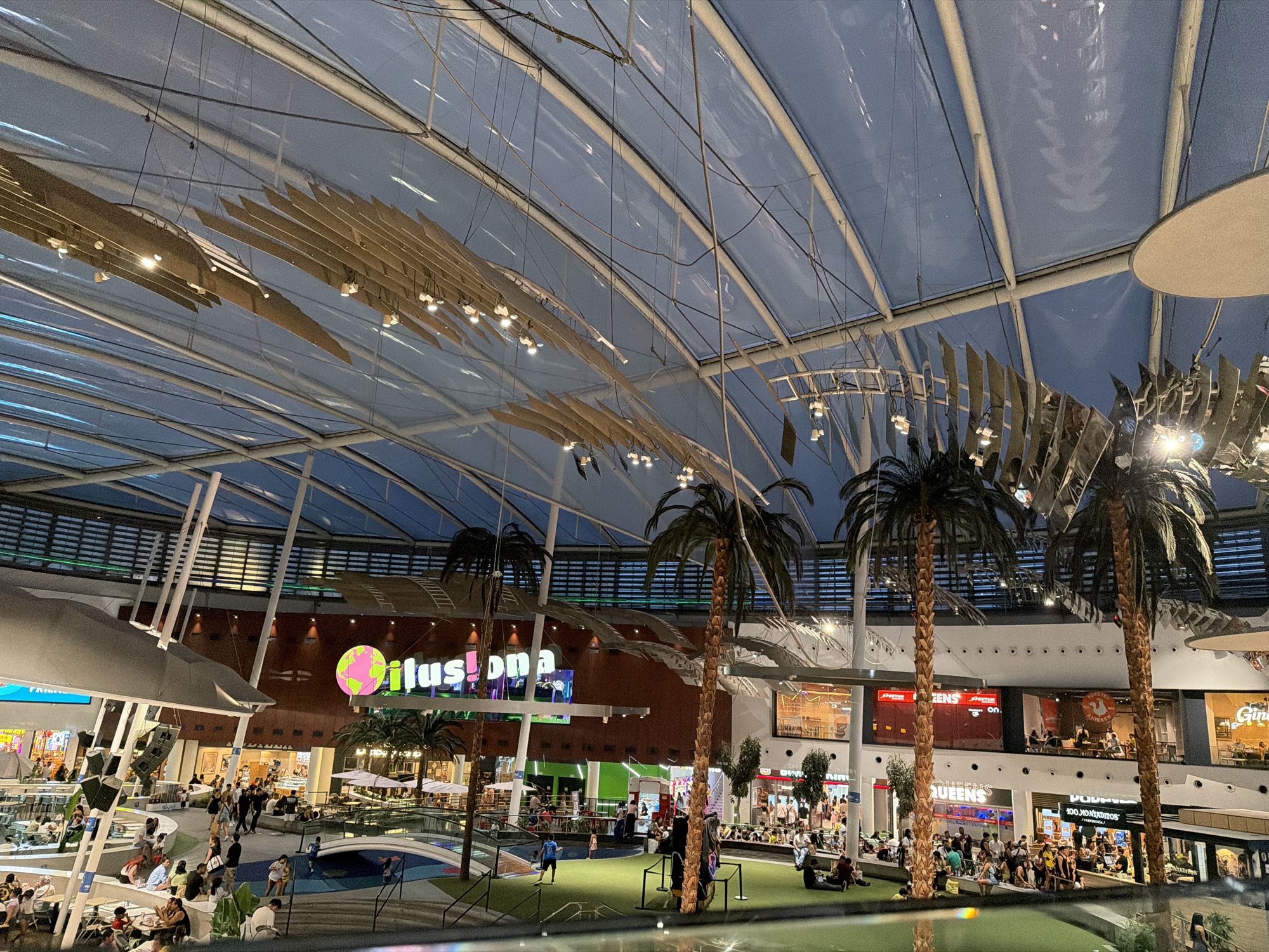 The image depicts the interior of a modern, multi-level shopping mall with a transparent, domed roof. The architecture features a spacious atrium, walkways, escalators, and various retail outlets. Several tall, artificial palm trees are interspersed throughout the mall, contributing to a tropical or resort-like ambiance. People are seen walking, sitting, and dining at various levels. The mall is illuminated by a combination of overhead lighting and the natural light filtering through the roof. A large, brightly colored sign that reads "ilusión" with a stylized globe is prominently visible on the upper level. The overall scene suggests a bustling commercial and recreational space.