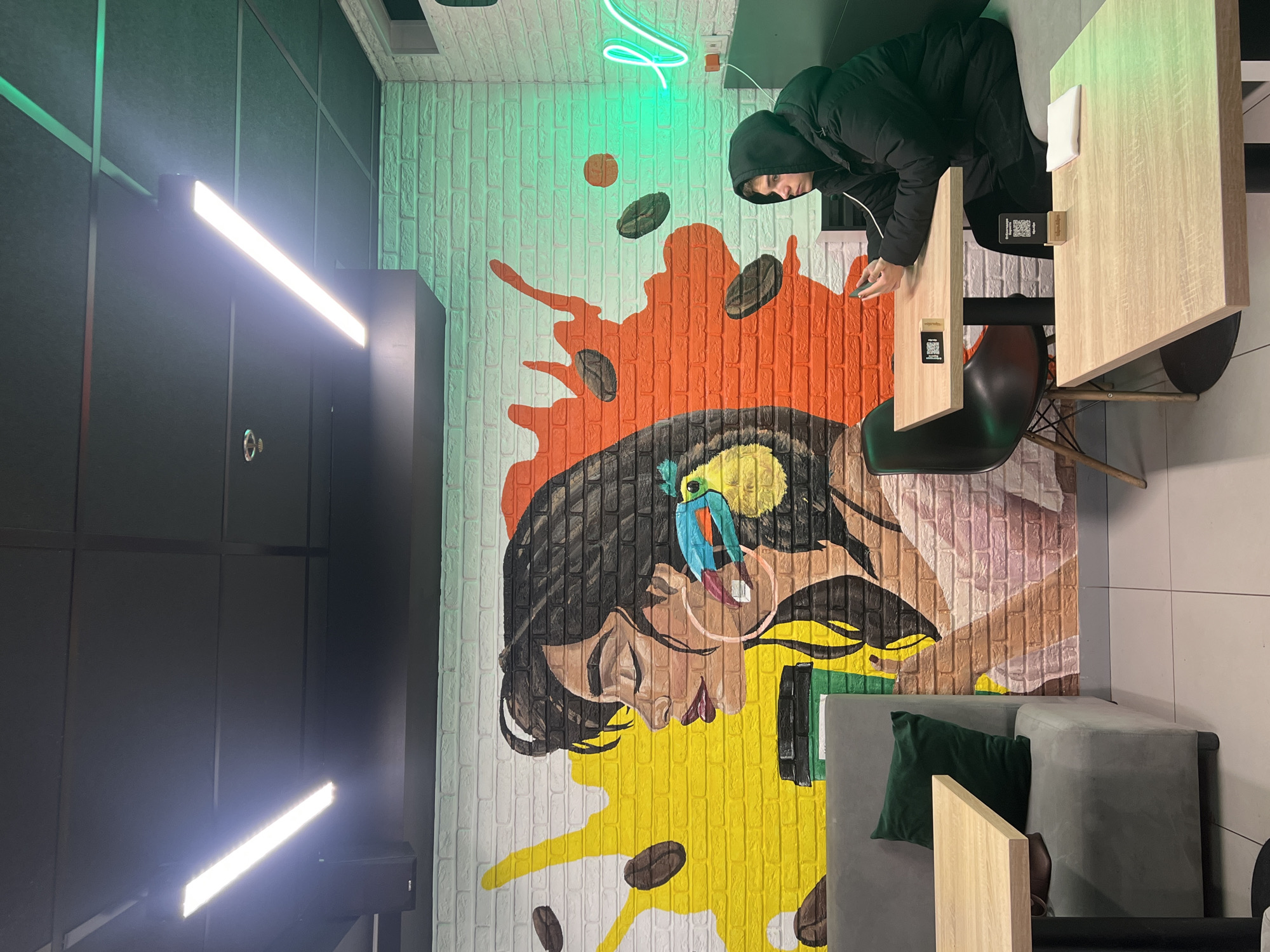 This image captures the interior of a modern establishment, likely a cafe or co-working space, in Saint Petersburg, Russia. The scene is dominated by a vibrant, multi-colored mural painted directly onto a white brick wall. The mural features a stylized portrait of a woman with dark hair, a small, colorful bird on her head, and splashes of yellow and orange paint, accompanied by scattered coffee beans.
A person, wearing a black hooded jacket, is seated at a light-colored wooden table on the right side of the frame. They are looking down at a smartphone in their hands, seemingly engaged in an activity on the device. Next to them is a black chair with a distinctive wire base. Another similar table is visible to the right, featuring a white napkin and a small stand with a QR code. More QR codes are visible on the tables, likely for menus or payment.
To the left, a grey sofa with a dark green throw pillow provides additional seating. The walls opposite the mural are dark grey or black, with a paneled design. A bright linear LED light fixture illuminates this dark wall. Above the mural, a bright green neon sign with an abstract design adds to the contemporary aesthetic. The overall lighting suggests an indoor setting, likely creating a cozy or trendy atmosphere.
