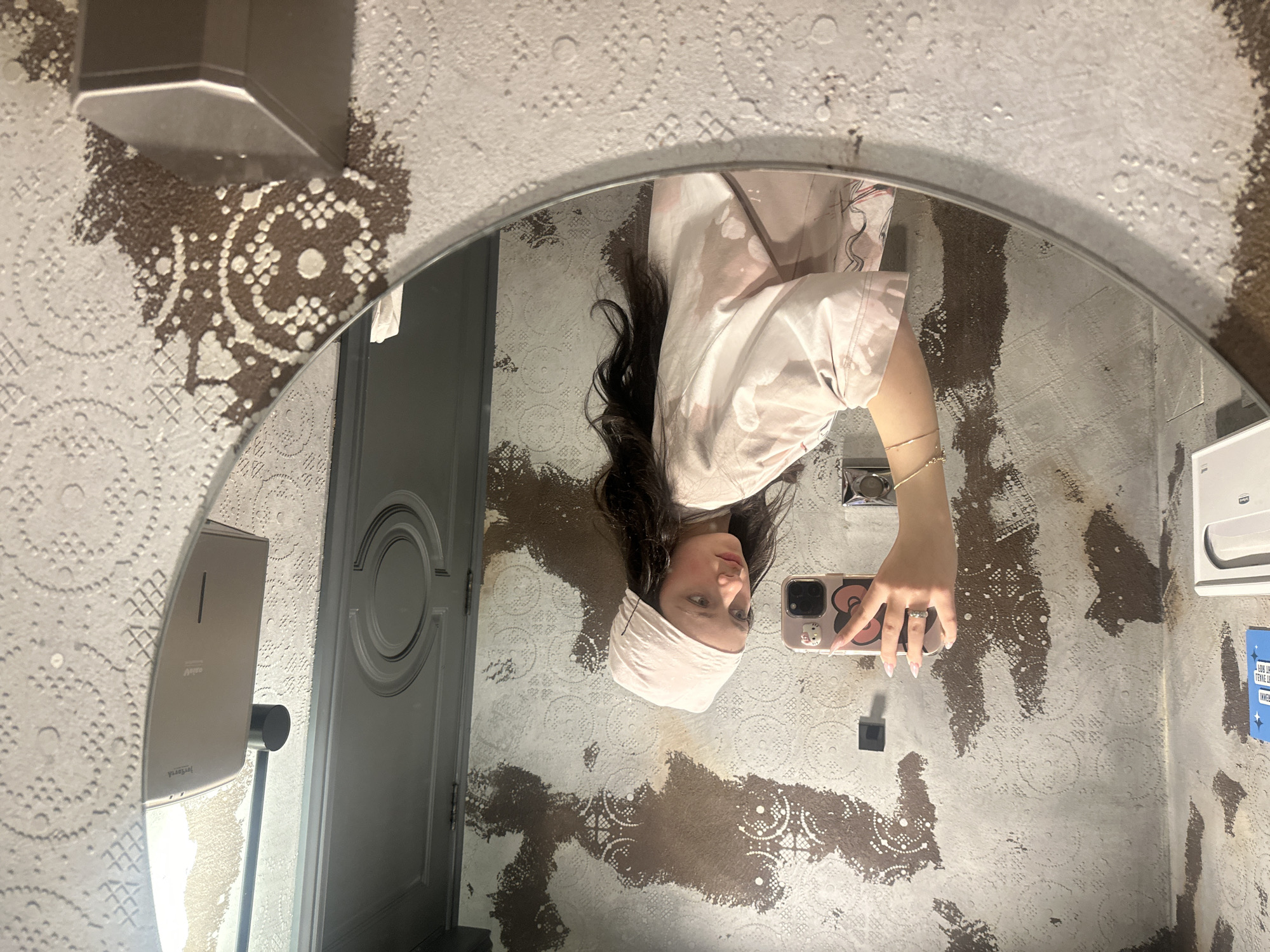 The image shows a person taking a selfie in a mirror. The background consists of a restroom with patterned wallpaper and various fixtures, including a soap dispenser, a door, and a paper towel dispenser. The person is wearing a light-colored head covering and clothing. The phone case is light pink with cartoon figures. The image was likely taken indoors. Without additional information, I can't confirm that this image was taken in Yerevan, Armenia.