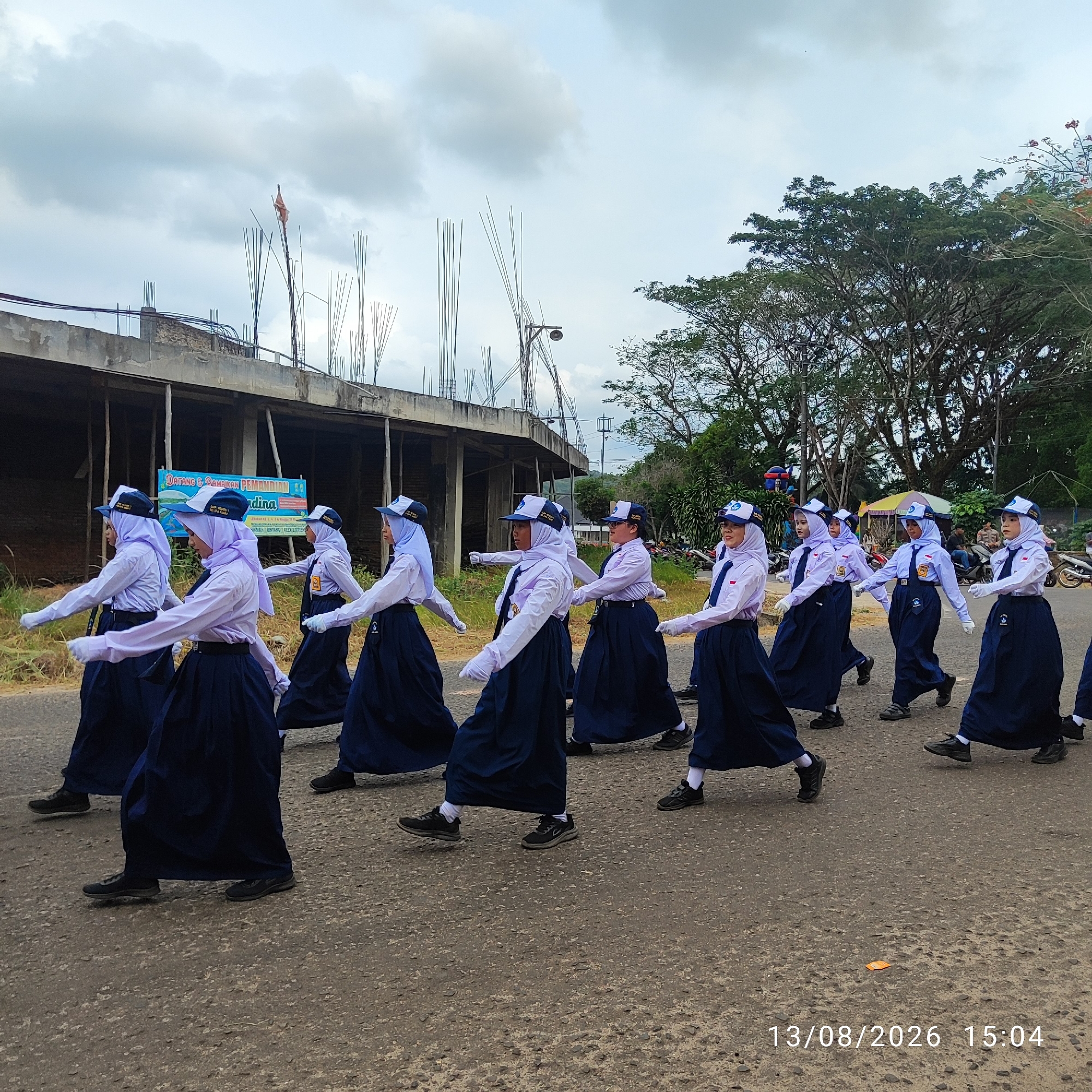 In Buding, Indonesia, on August 13, 2026, at 3:04 PM, a group of Indonesian schoolchildren, likely participating in a parade, marches in single file down a paved road. The children are wearing matching uniforms: white long-sleeved shirts, light purple hijabs, dark blue pleated skirts, and dark blue caps. They are also wearing white gloves and dark shoes. The parade is taking place outdoors, with a cloudy sky overhead.
In the background, there is a partially constructed building with visible rebar and scaffolding. A banner is hanging from the building with Indonesian text. To the right of the road, there is a dense line of trees. The overall scene suggests a community event or school activity occurring in an semi-urban or rural setting in Buding.