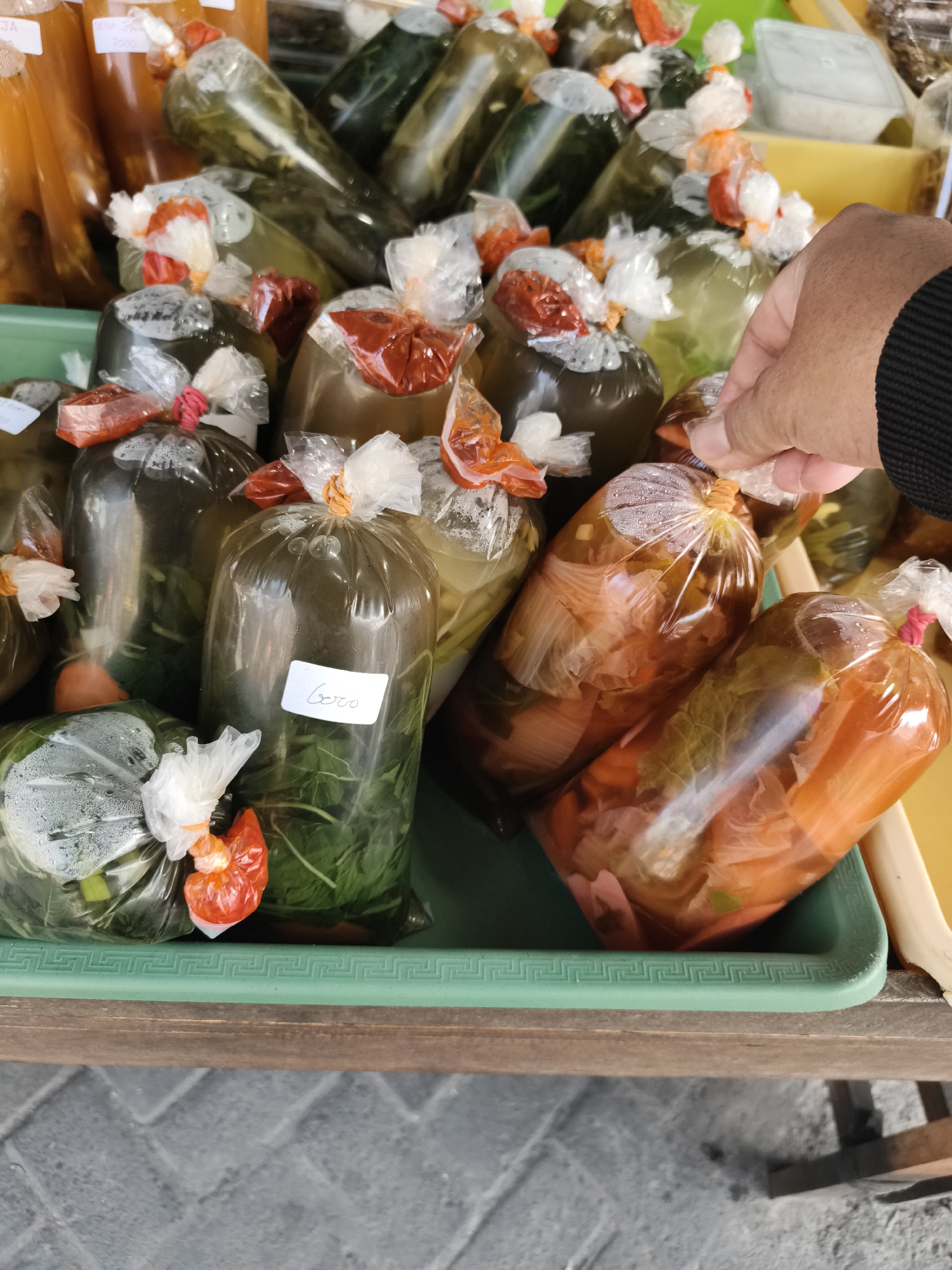 In Petiken, Indonesia, a person's hand is reaching for a small, clear plastic bag containing food. The bag is tied with an orange string and appears to contain vegetables. This bag is part of a larger collection of similarly packaged food items, some filled with leafy green vegetables and others with ingredients that appear to be pickled vegetables like carrots and cabbage, all displayed in a green plastic tray. The background shows more bags of food, some with orange-colored contents, and hints of a yellow surface and a gray tiled ground. The scene suggests a market or food stall setting.